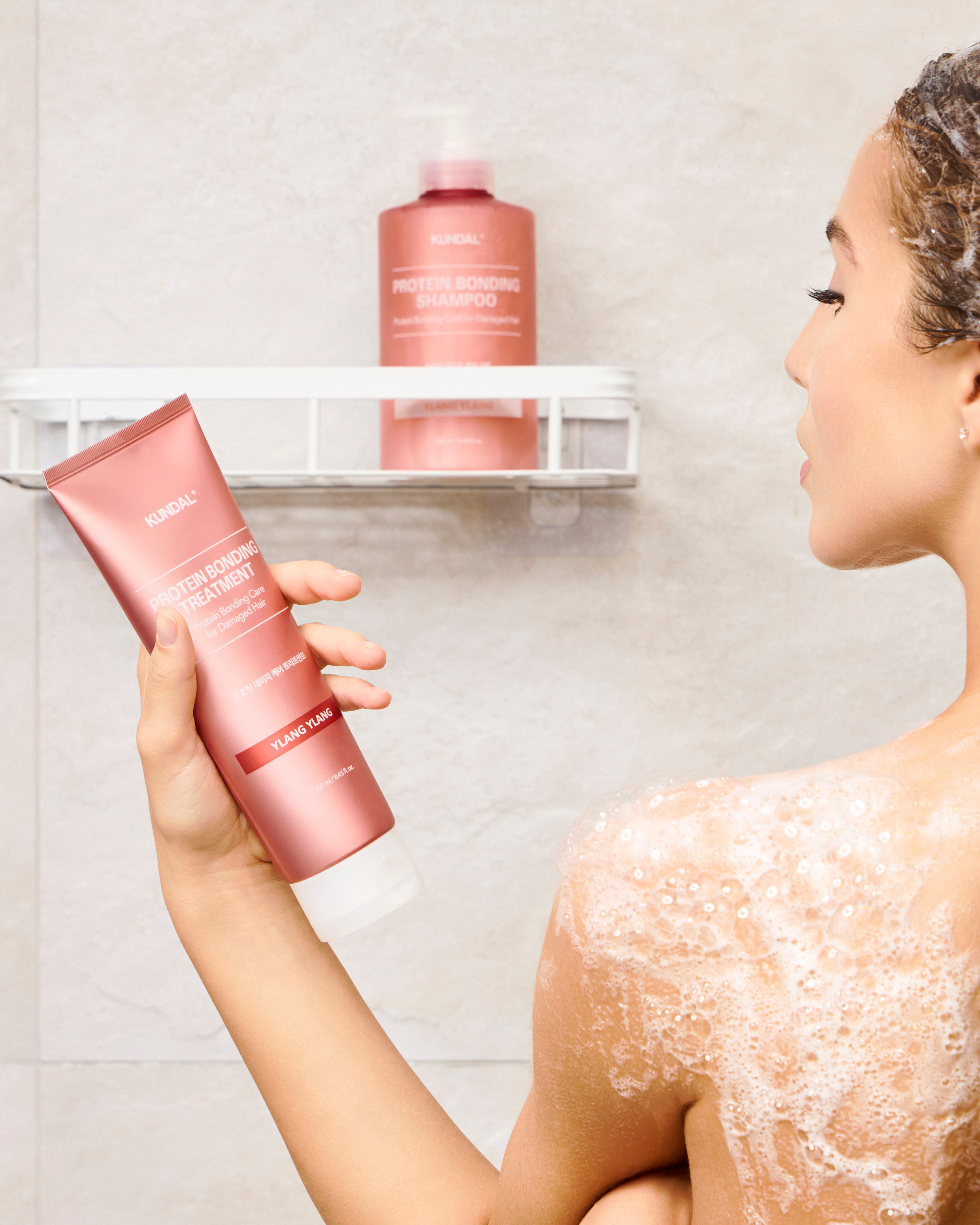 Eine Frau unter der Dusche, mit Schaum auf Schulter und Haaren, hält eine rosa Tube "Kundal Protein Bonding Treatment" in der Hand. Im Hintergrund steht eine "Kundal Protein Bonding Shampoo" Flasche.