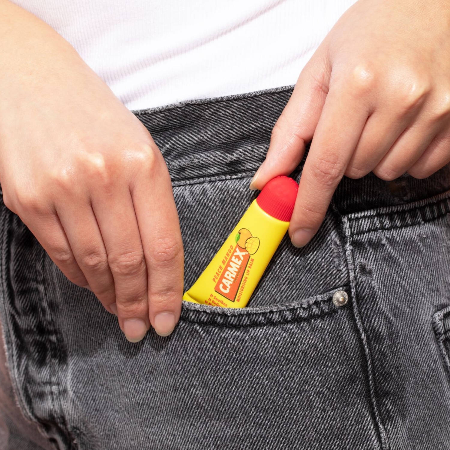Gelbe Tube CARMEX Peach Mango Lippenbalsam in grauer Jeanshosentasche. Rote Kappe sichtbar. Hände greifen zur Tube.