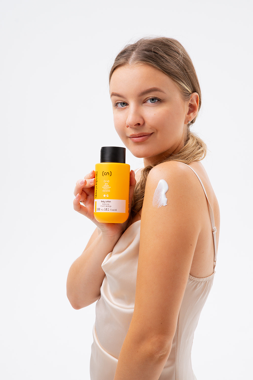 Frau hält gelbe SKINCYCLOPEDIA Glow Body Lotion. Lotion auf Arm, für strahlende Haut.