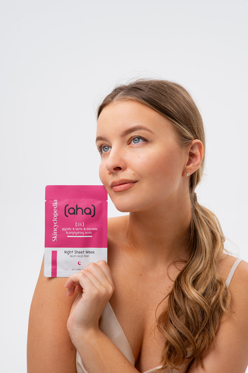Porträt einer Frau, die eine SKINCYCLOPEDIA AHA/PHA Sheet Maske in der Hand hält. Die Maske ist pink-weiß und bewirbt ein sanftes Peeling.