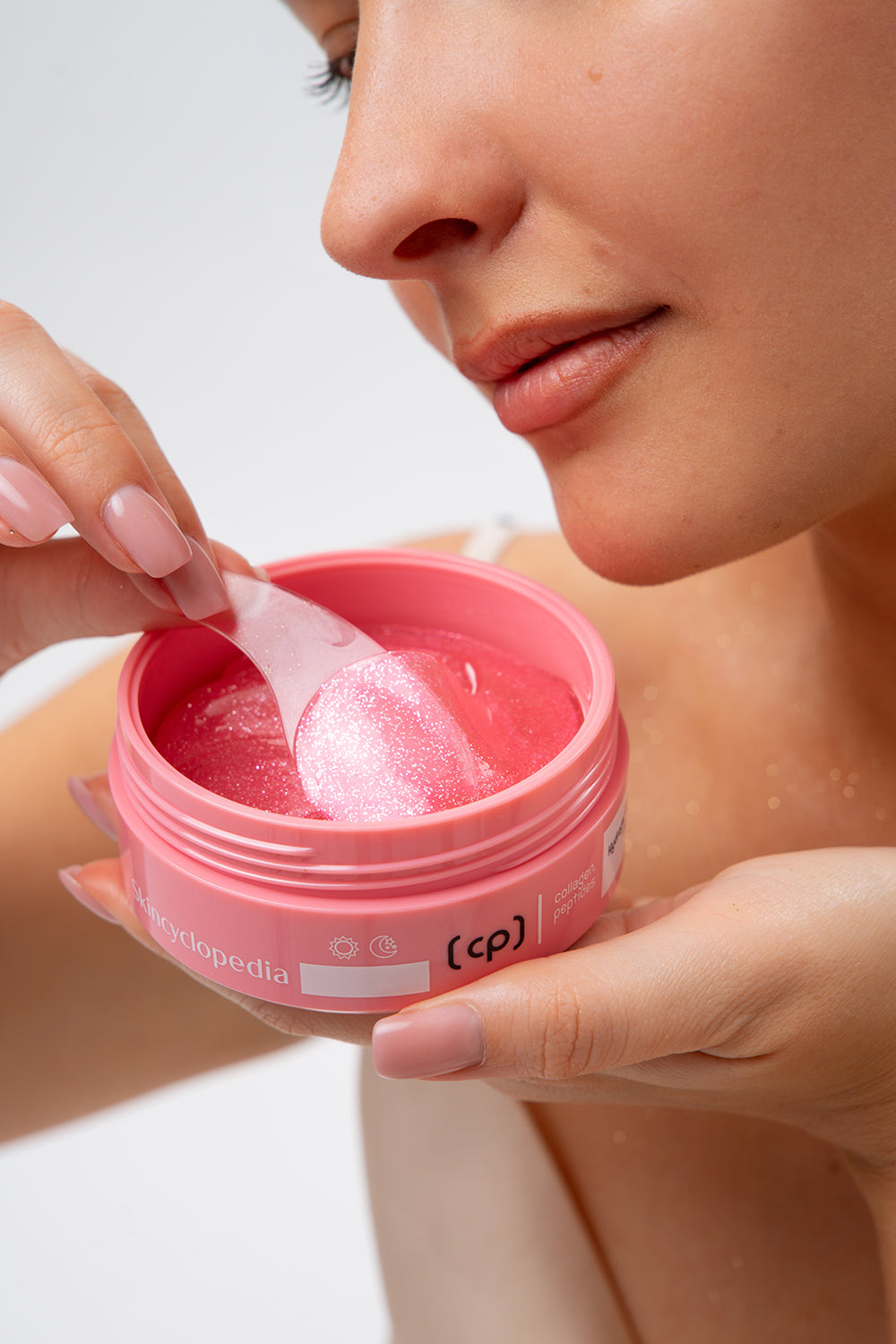 Frau entnimmt mit Spatel ein Collagen Augenpatch aus einer pinken Dose. Dose mit Aufschrift "Skincyclopedia". Inhalt glitzert.