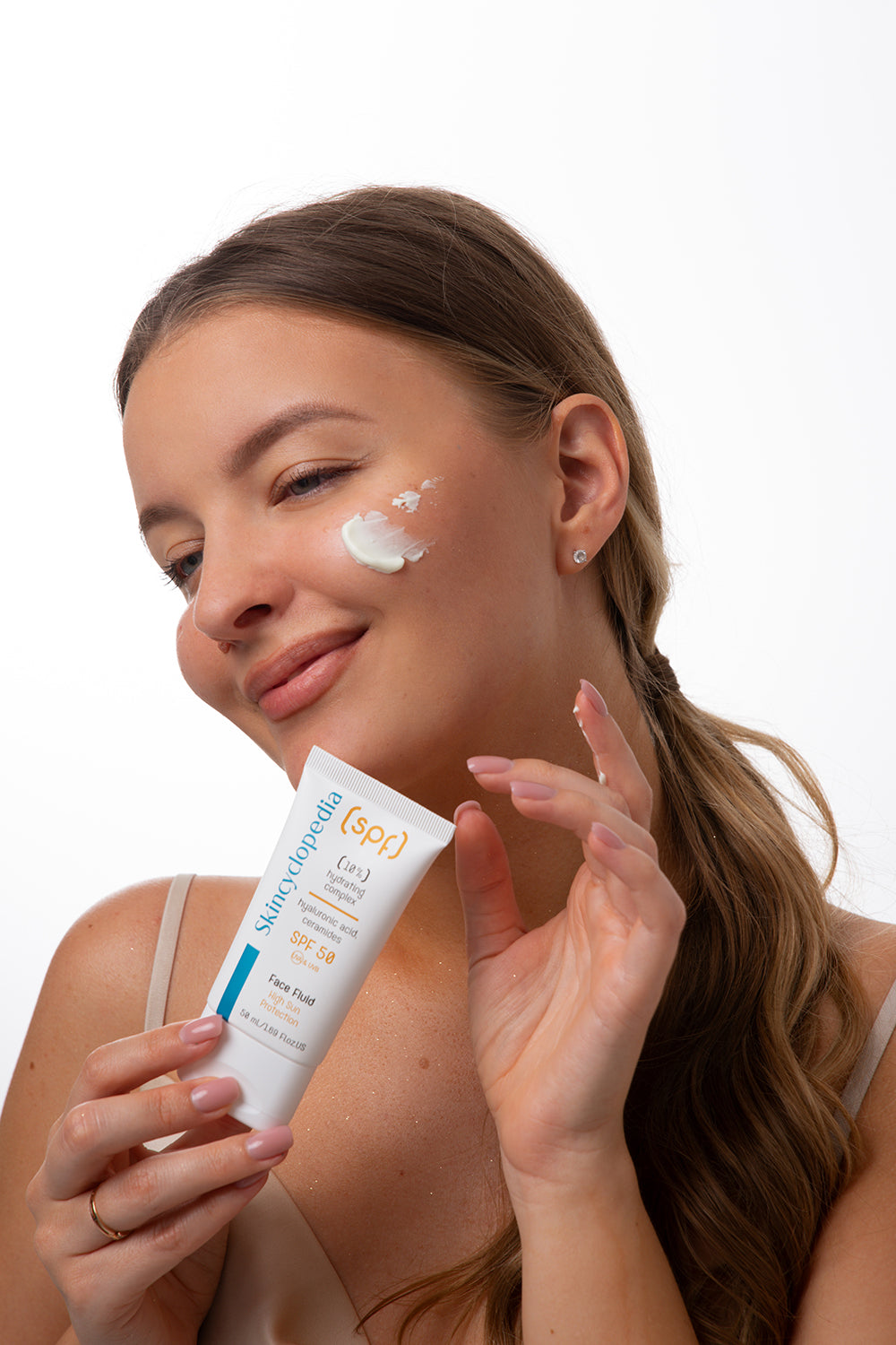 Frau mit SKINCYCLOPEDIA SPF 50 Fluid und einem Klecks Creme auf der Wange. Das Produkt verspricht Feuchtigkeit und UV-Schutz.
