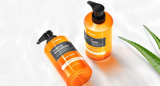 Zwei honigfarbene Flaschen mit schwarzem Pumpspender, beschriftet mit "Honey & Macadamia Body Wash", liegen auf weißem Stoff. Daneben Aloe Vera Blätter.