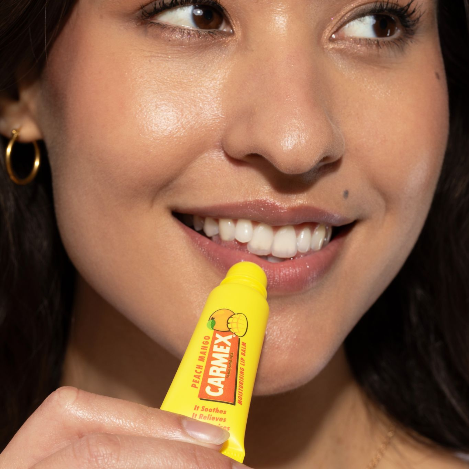 Lächelnde Frau trägt Carmex Tube Peach Mango Lippenbalsam auf. Fokus auf gelbe Tube mit Pfirsich und Mango Design. Gepflegte Lippen.