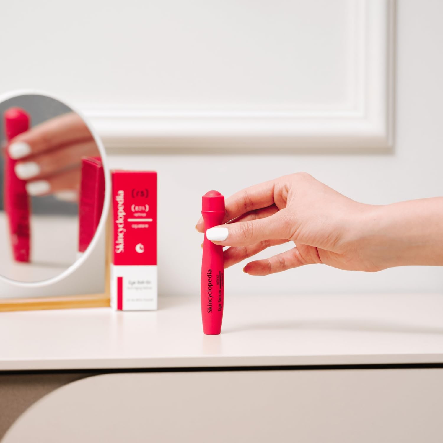 Uma mão segura o roll-on de retinol SKINCYCLOPEDIA cor-de-rosa numa penteadeira, ao lado do espelho e da embalagem