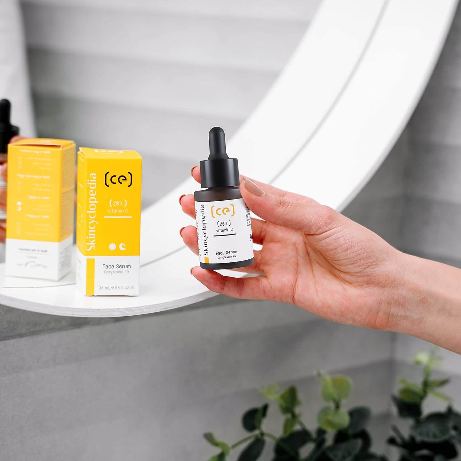 Hand hält Vitamin C 20% Serum-Fläschchen. Gelbe Produktverpackung und Flasche stehen auf einem Regal.