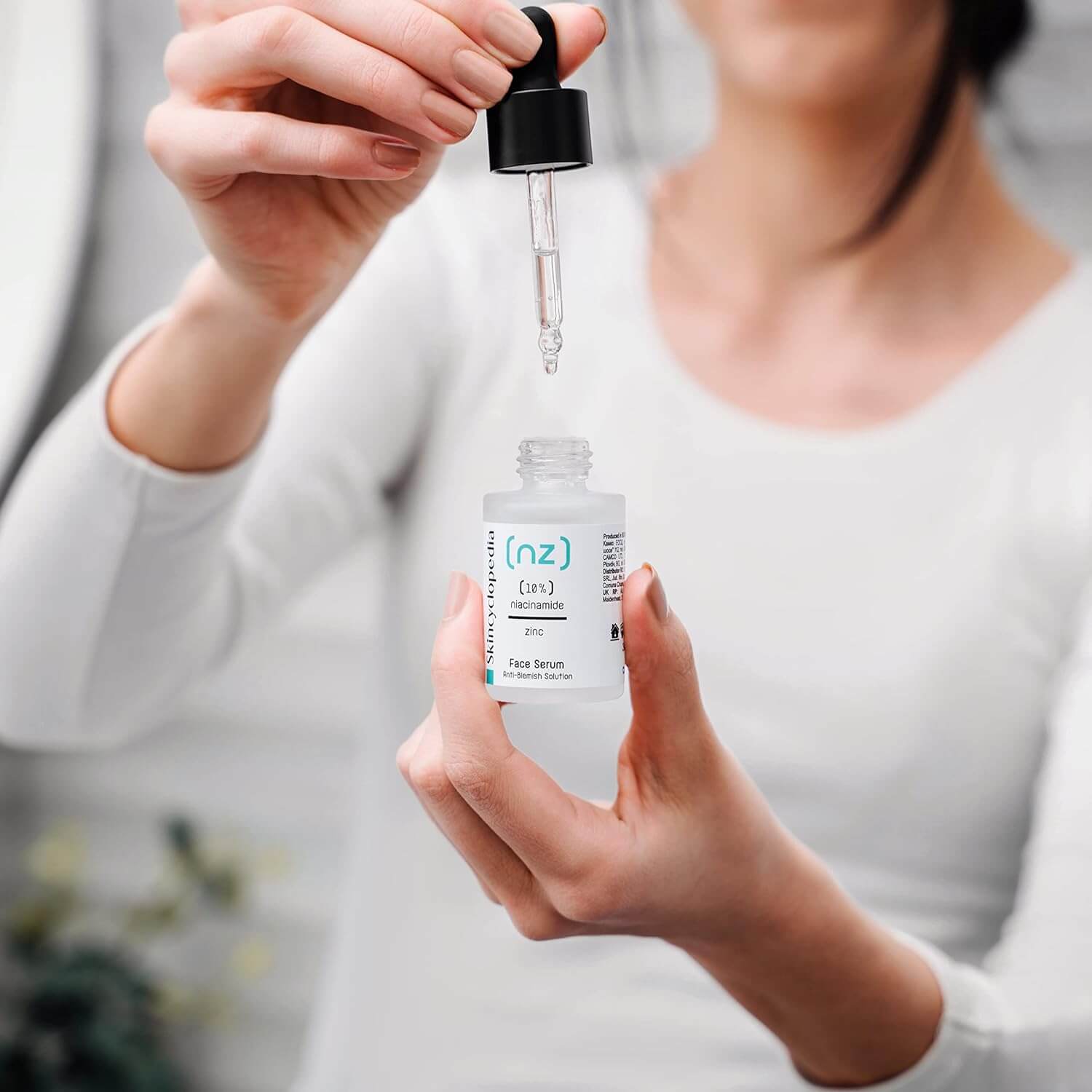 Eine Person hält das SKINCYCLOPEDIA Niacinamide 10 % Serum und eine Pipette. Ein Tropfen Serum fällt in die geöffnete Flasche.