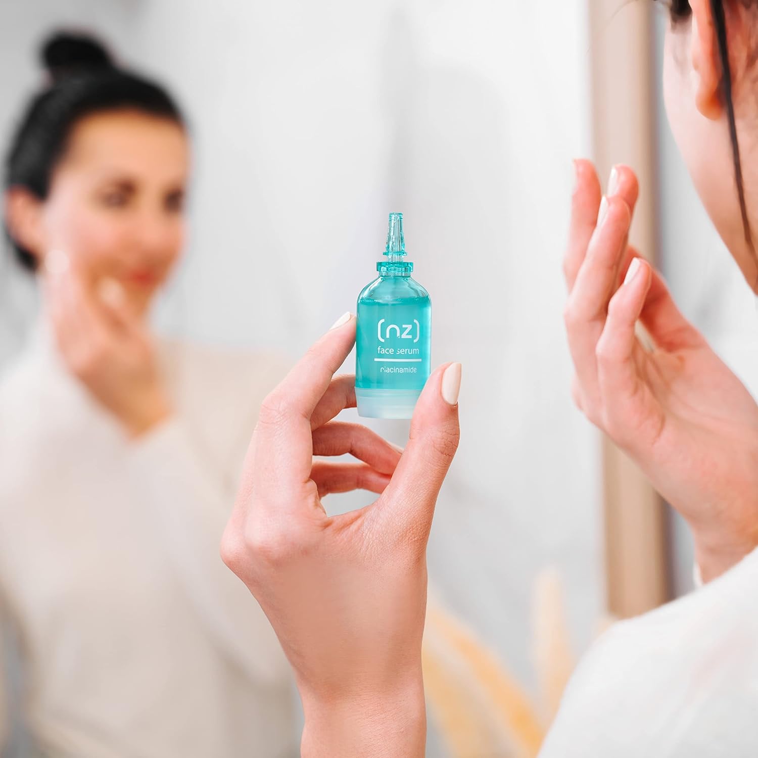 Eine Frau hält ein blaues Gesichtsserum in der Hand, während sie in den Spiegel schaut. Das Serum hat eine klare Flüssigkeit und die Aufschrift "(nz) face serum niacinamide".