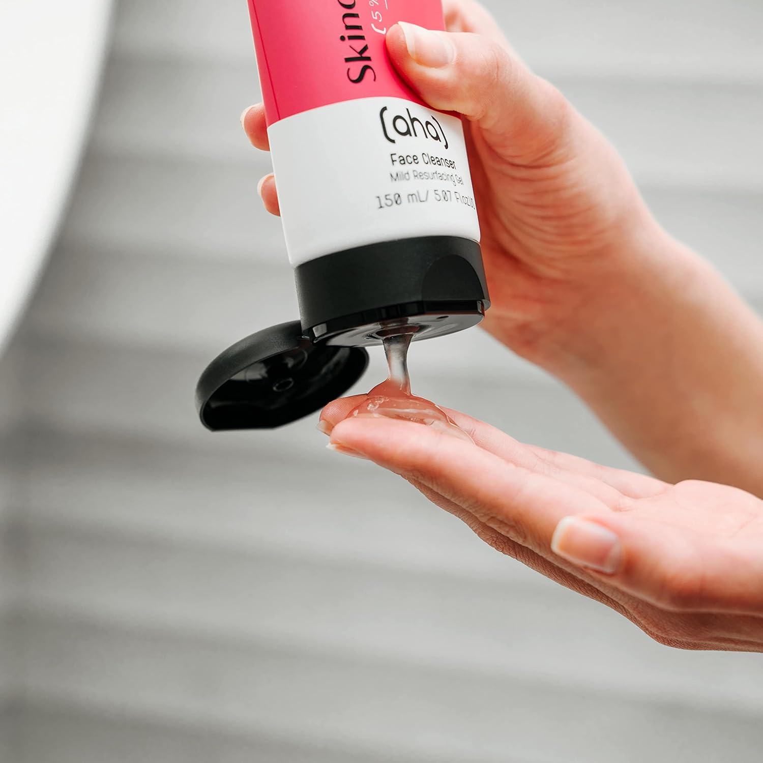 Hand giesst AHA/PHA Gesichtsreiniger auf andere Hand. Gelartige Textur sichtbar. Tube in Pink, Weiss, Schwarz. Klärend & Peelend.