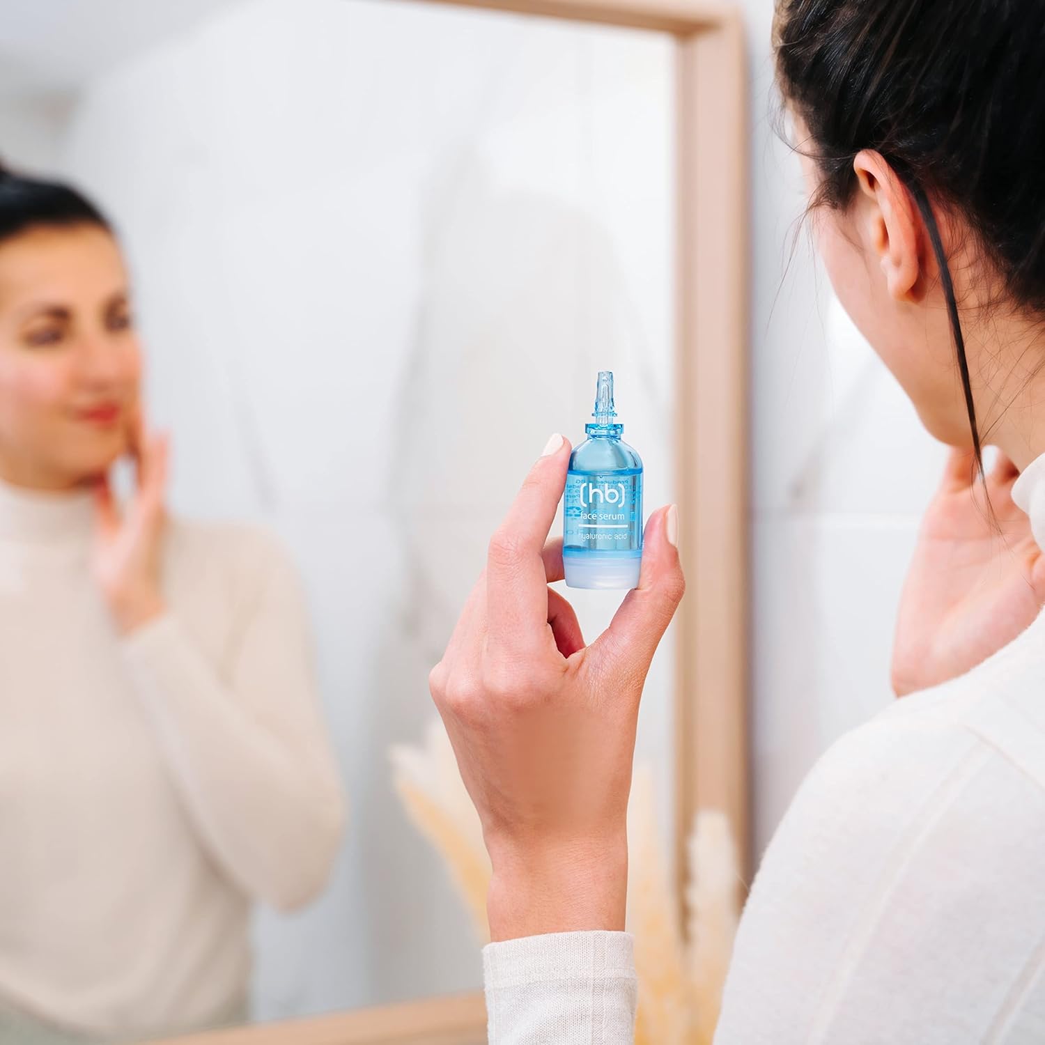 Eine Frau hält ein SKINCYCLOPEDIA Hyaluron Serum vor einem Spiegel. Im Spiegelbild ist die Frau mit Serum auf ihren Wangen zu sehen.