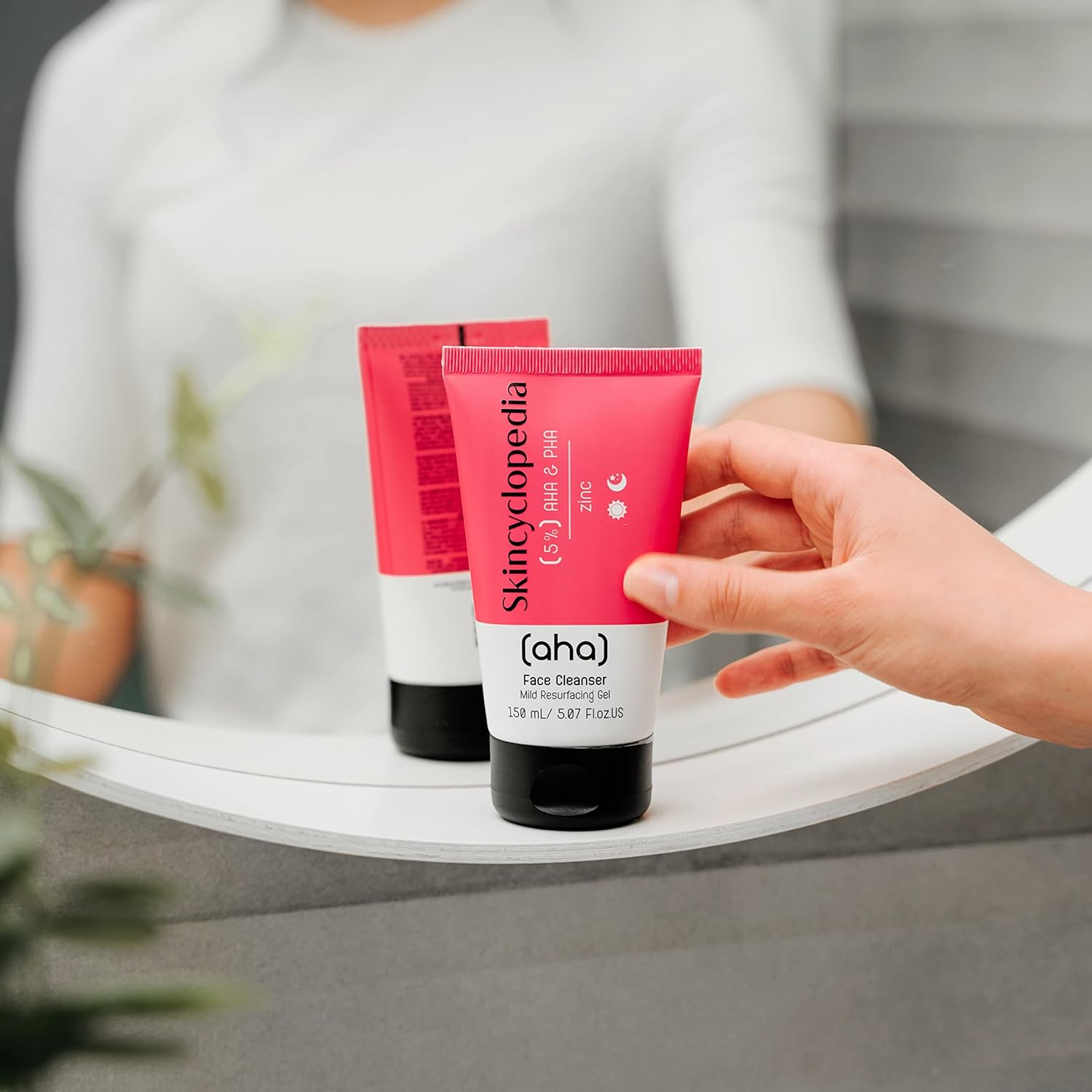 Nahaufnahme von zwei SKINCYCLOPEDIA AHA/PHA Gesichtsreiniger Tuben in pink und schwarz. Eine Hand hält eine der Tuben vor einem Spiegel.