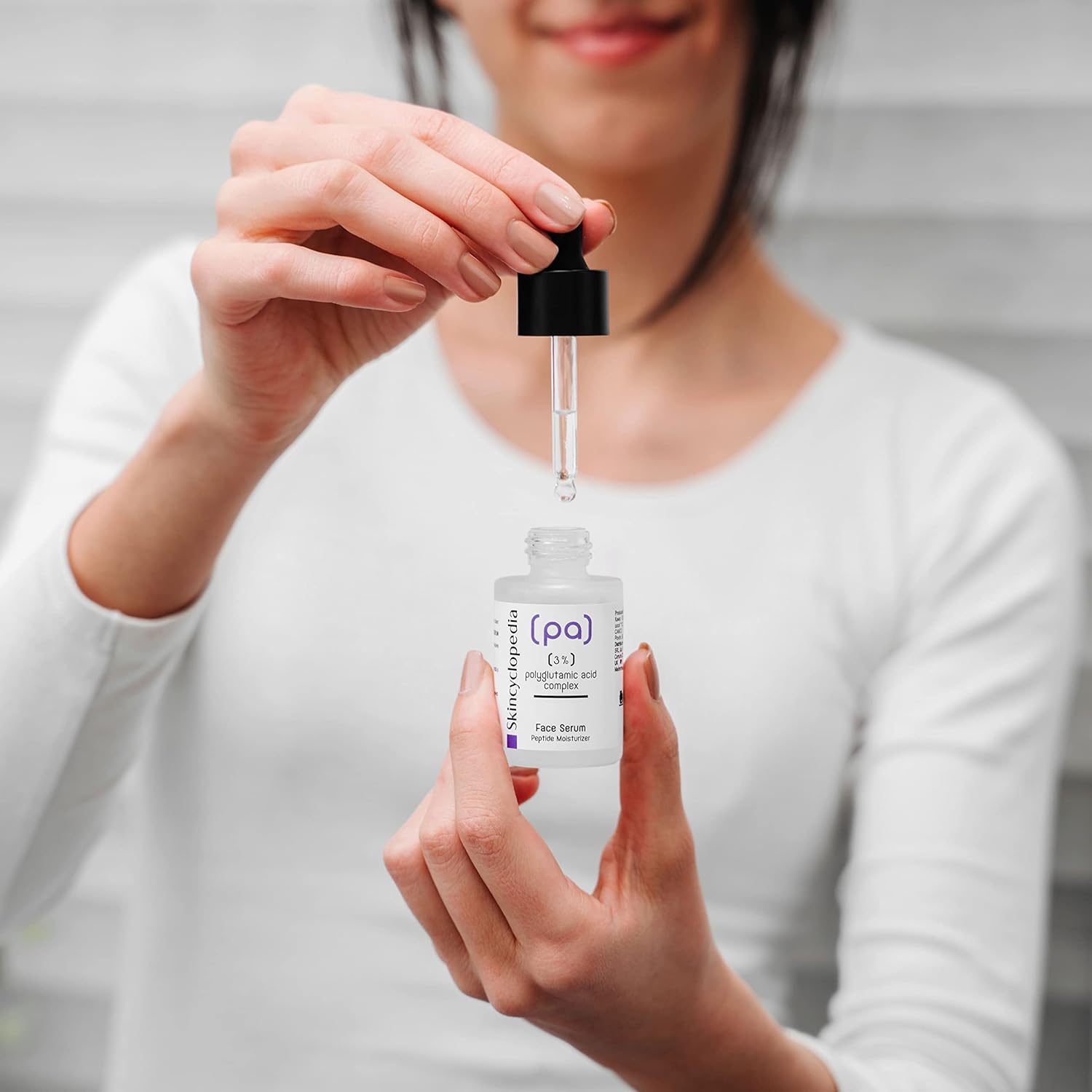 Eine Person hält ein SKINCYCLOPEDIA Polyglutaminsäure Serum mit Pipette in der Hand. Serumtropfen fallen. Fokus auf das Feuchtigkeitsserum.