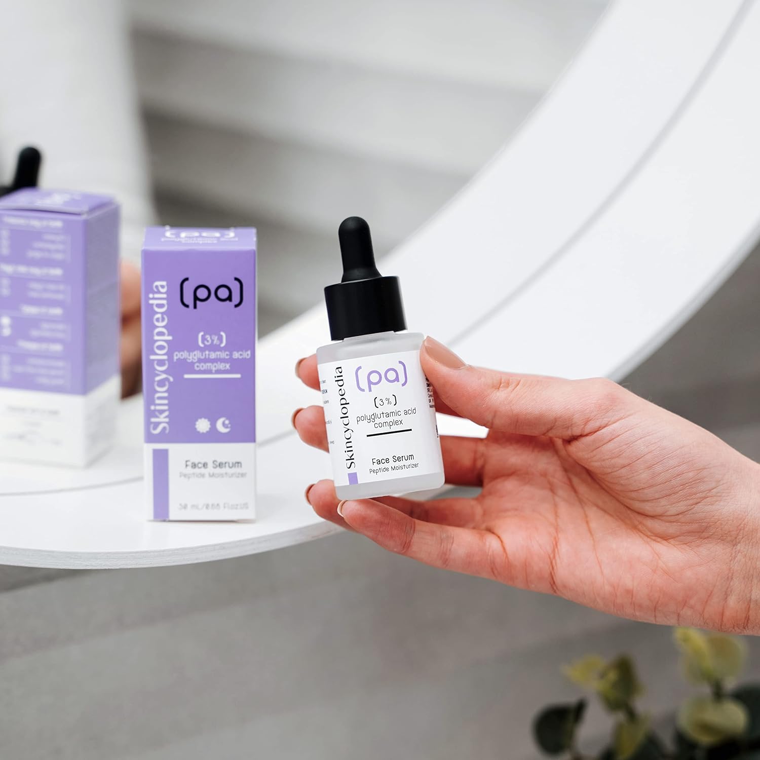 Eine Hand hält ein SKINCYCLOPEDIA Polyglutaminsäure Serum. Eine Packung des Serums steht daneben. Im Hintergrund ist ein Spiegel.
