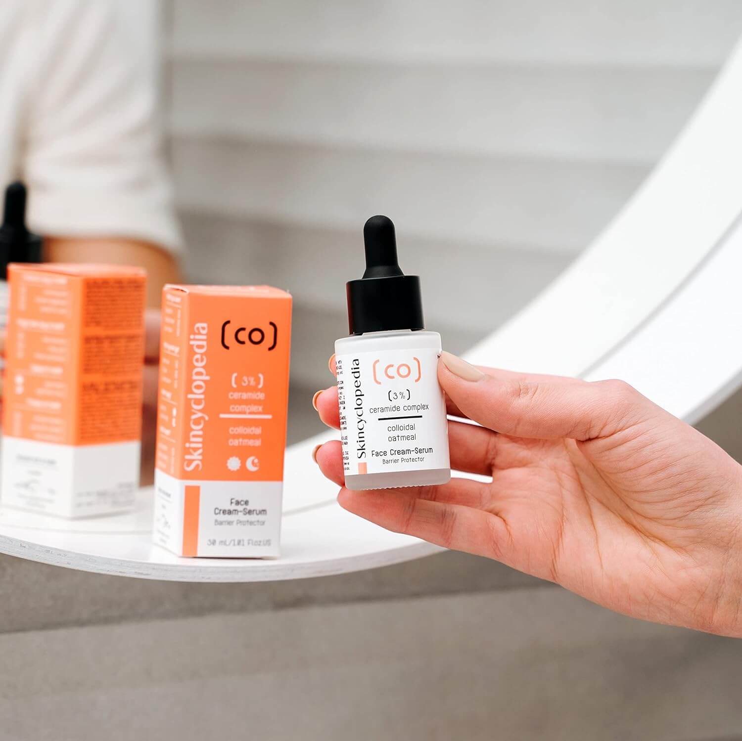 Nahaufnahme: Hand hält SKINCYCLOPEDIA Ceramide Serum mit Pipette. Im Hintergrund Produktverpackung in Orange-Weiss. Fokus auf Serum, stärkt & beruhigt die Haut.
