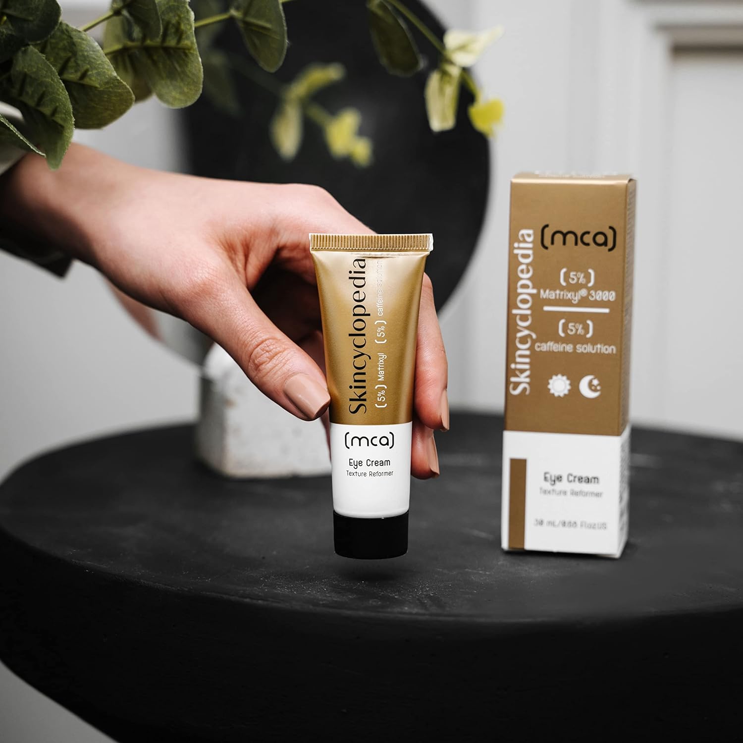 Uma mão segura o tubo branco e dourado do creme para os olhos SKINCYCLOPEDIA – Matrixyl & Cafeína 15 ml ao lado da sua embalagem