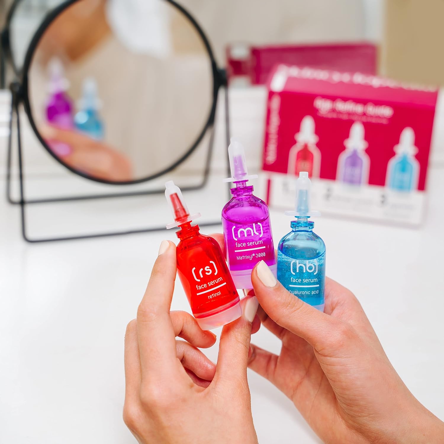 Nahaufnahme: Hände halten drei farbige Seren (Retinol, Matrixyl, Hyaluron) aus dem SKINCYCLOPEDIA Age Perfect Guide Set. Im Hintergrund ein Spiegel und die Produktverpackung.