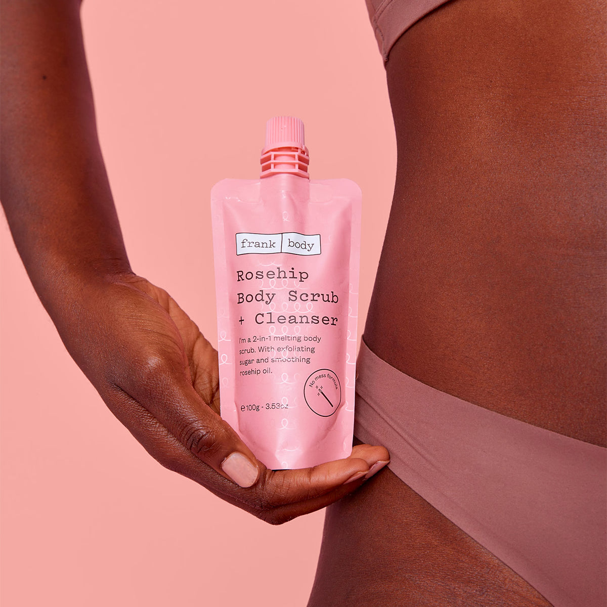Eine Hand hält den FRANK BODY Rosehip Body Scrub + Cleanser (ON-THE-GO) 100g Beutel vor rosafarbenem Hintergrund