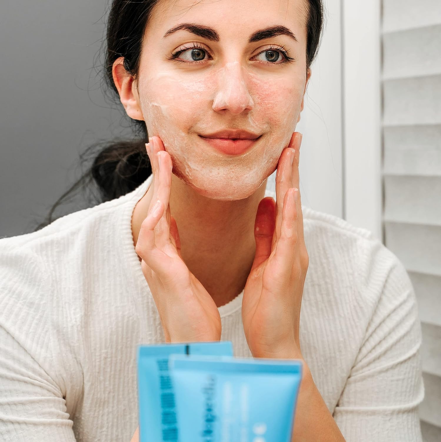 Frau massiert SKINCYCLOPEDIA Gesichtsreiniger mit 5% Feuchtigkeitskomplex sanft in die Gesichtshaut ein. Blaue Tube im Vordergrund.