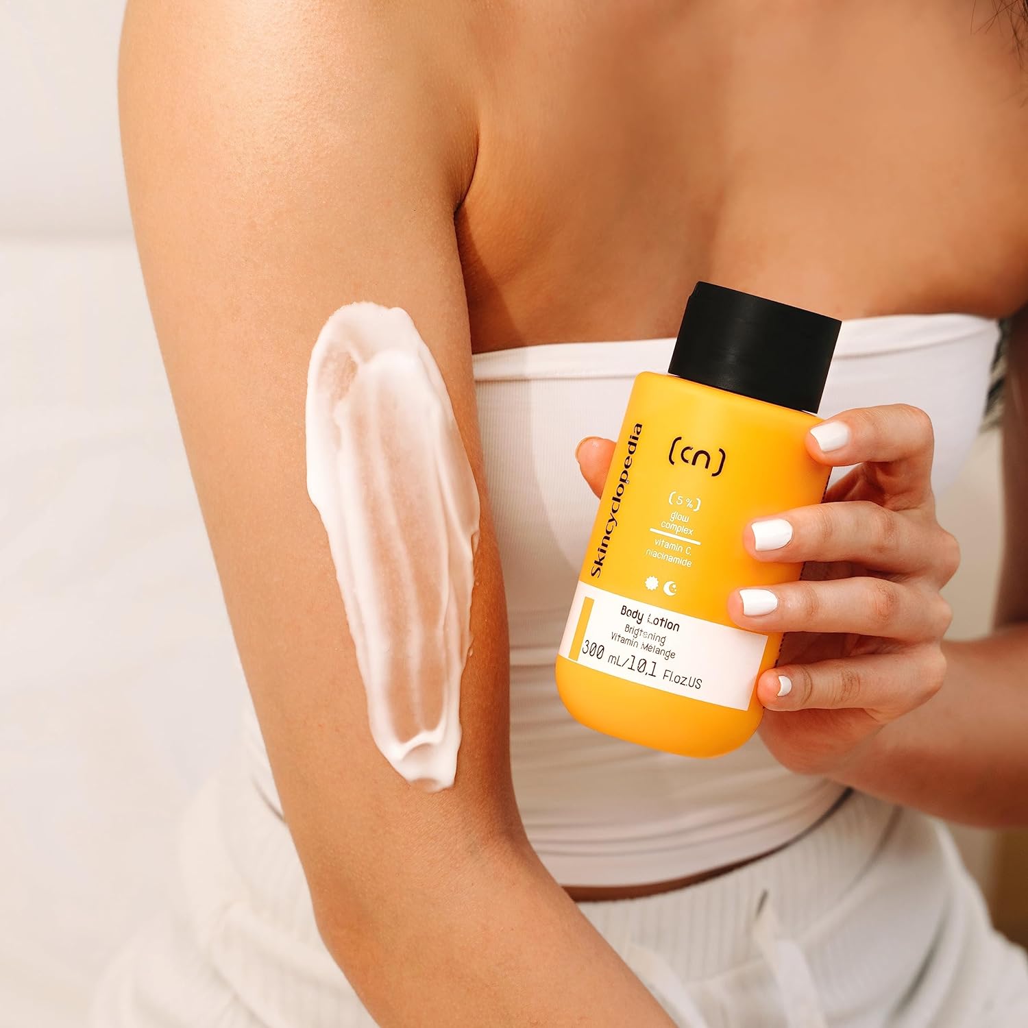 Eine Person cremt ihren Arm mit SKINCYCLOPEDIA Body Lotion ein. Die gelbe Flasche wird in der Hand gehalten. Sichtbar ist der 5% Glow Komplex Hinweis auf der Lotion.