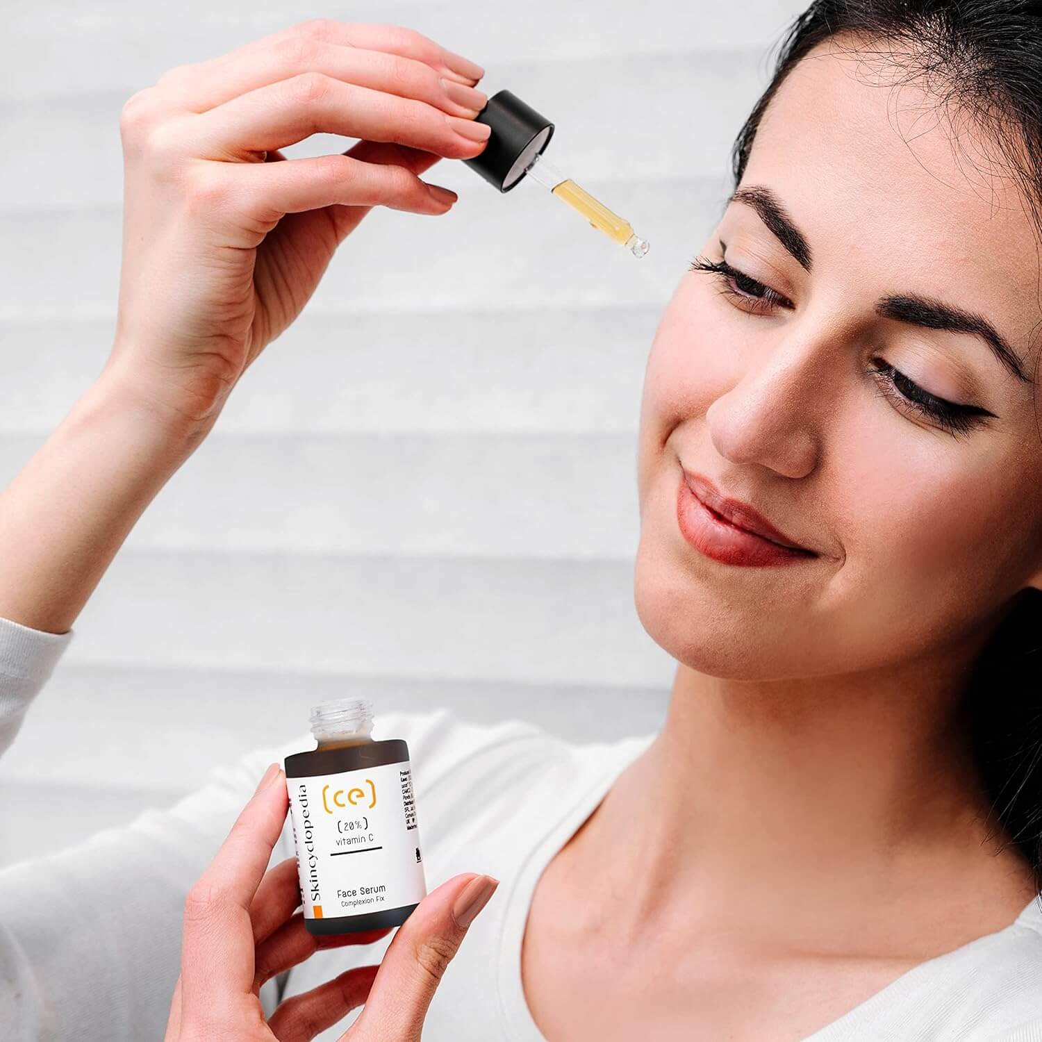 Frau mit Pipette und SKINCYCLOPEDIA Vitamin C 20% Serum. Sie hält die Pipette über ihr Gesicht. Fokus auf Serum-Flasche und Frau.