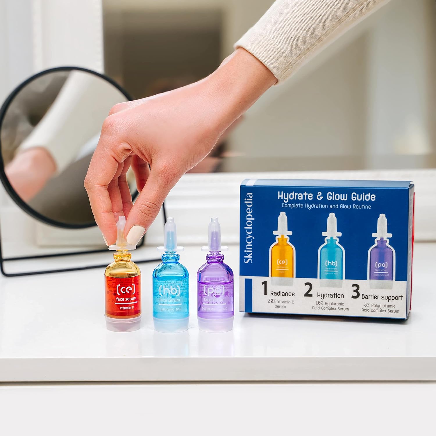 Das Bild zeigt das SKINCYCLOPEDIA Hydrate Guide 3er-Set. Die Hand einer Person hält das Vitamin C Serum. Im Hintergrund ist die Verpackung des Sets zu sehen.