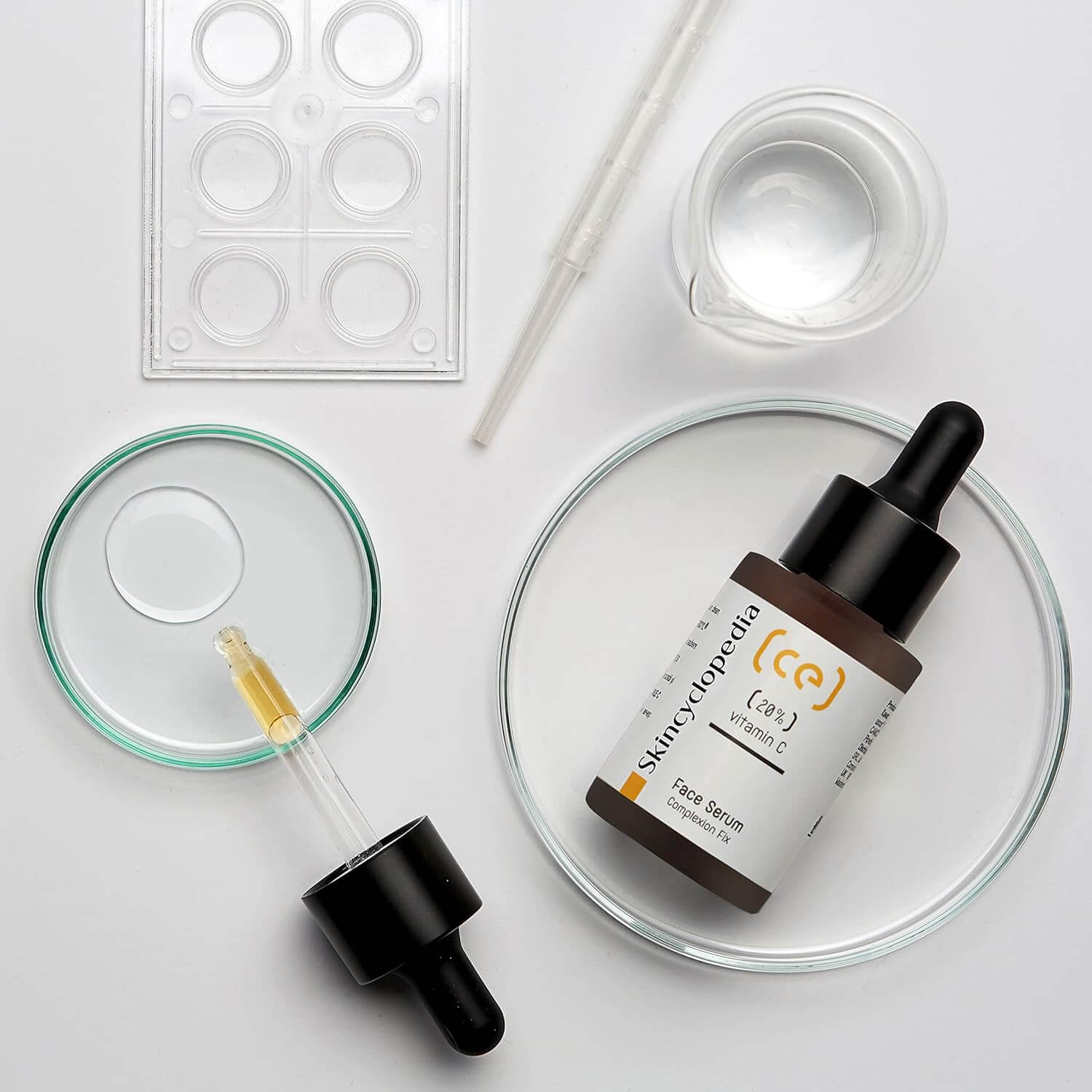 Ein braunes SKINCYCLOPEDIA Vitamin C 20 % Serum, Pipetten, Schalen und Messbecher auf weißem Hintergrund.