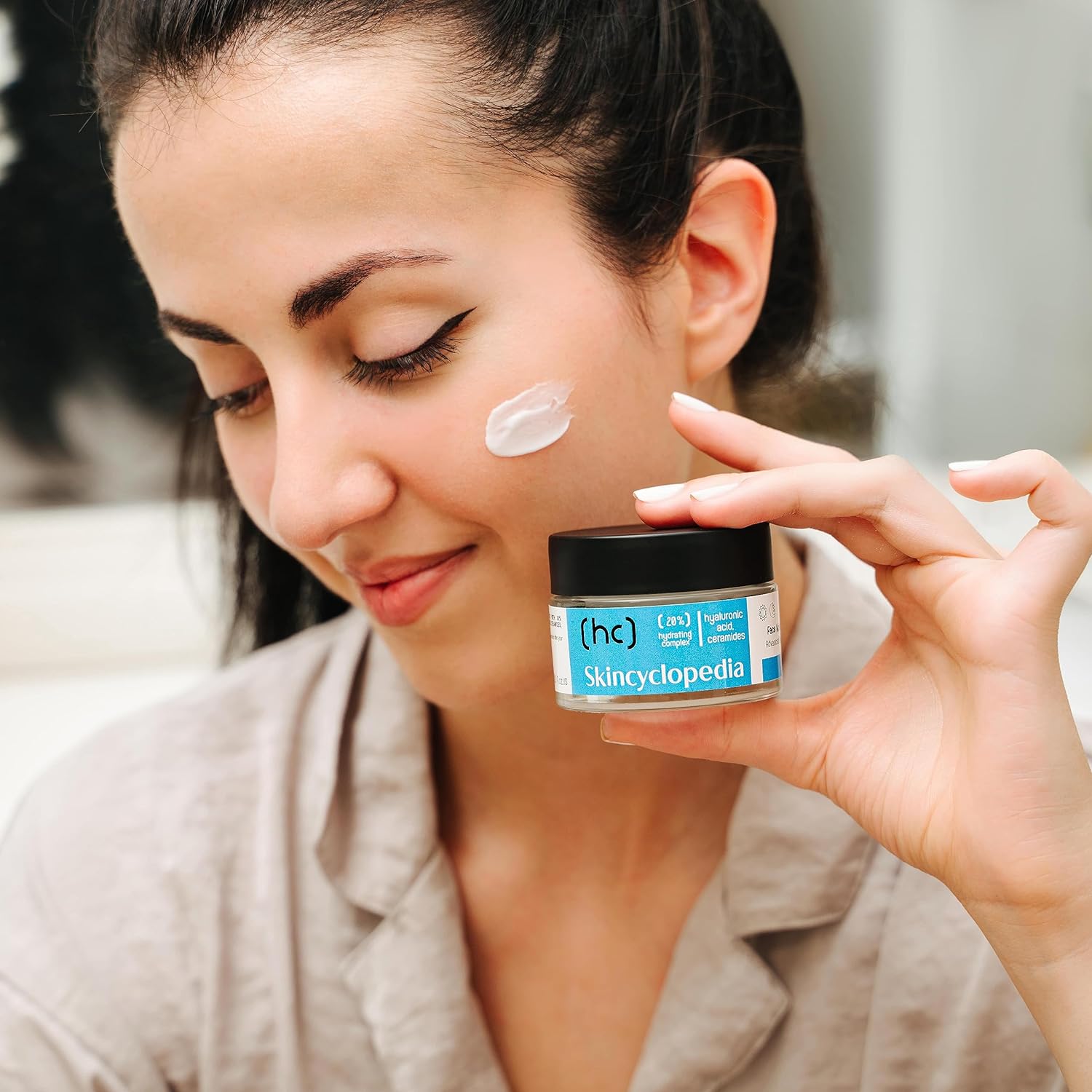 Frau mit Feuchtigkeitscreme auf Wange hält SKINCYCLOPEDIA Creme. Fokus auf das Produkt und die Anwendung. Intensive Hydration im Alltag.