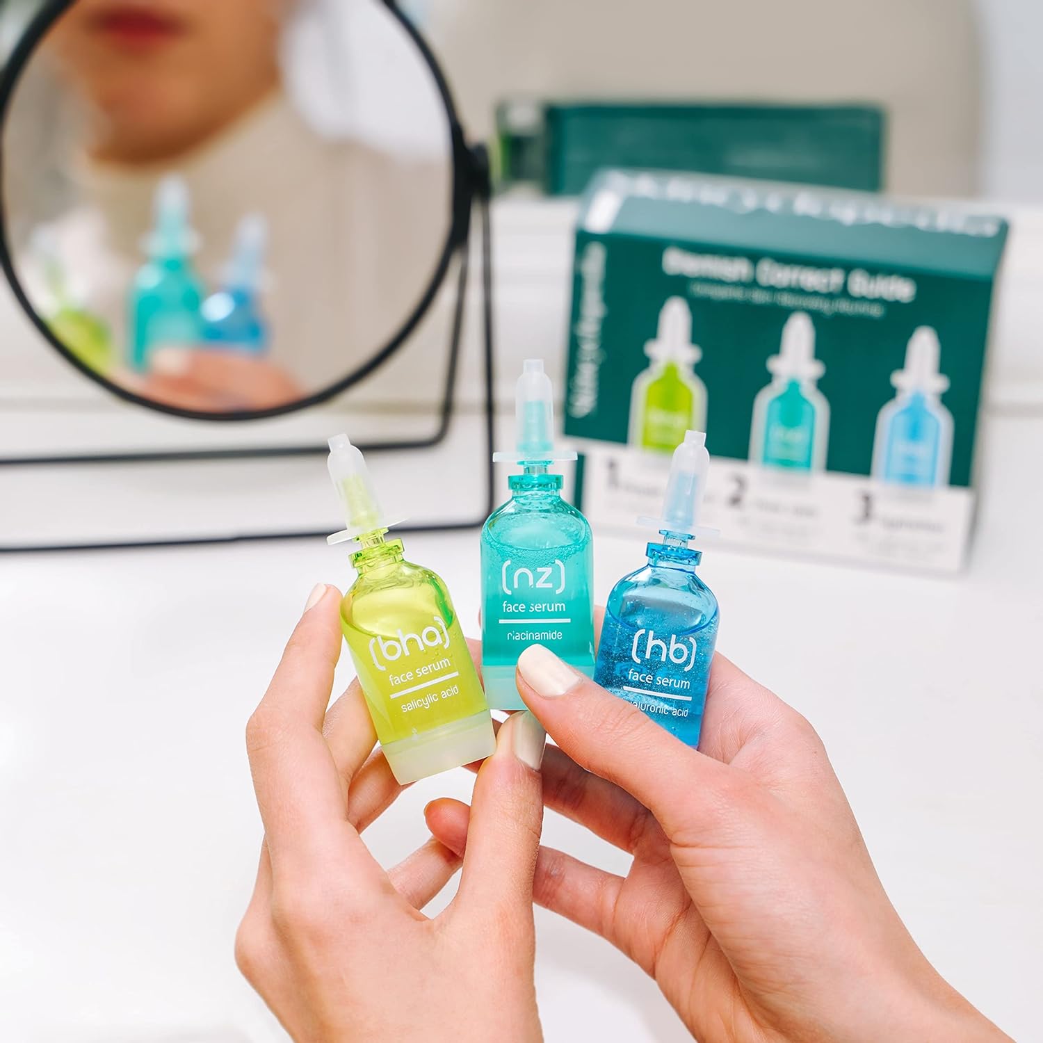 Zwei Hände halten drei bunte Gesichtsseren mit Pipettenverschlüssen, im Hintergrund ein Spiegel und eine Produktbox. Die Seren sind grün, türkis und blau.
