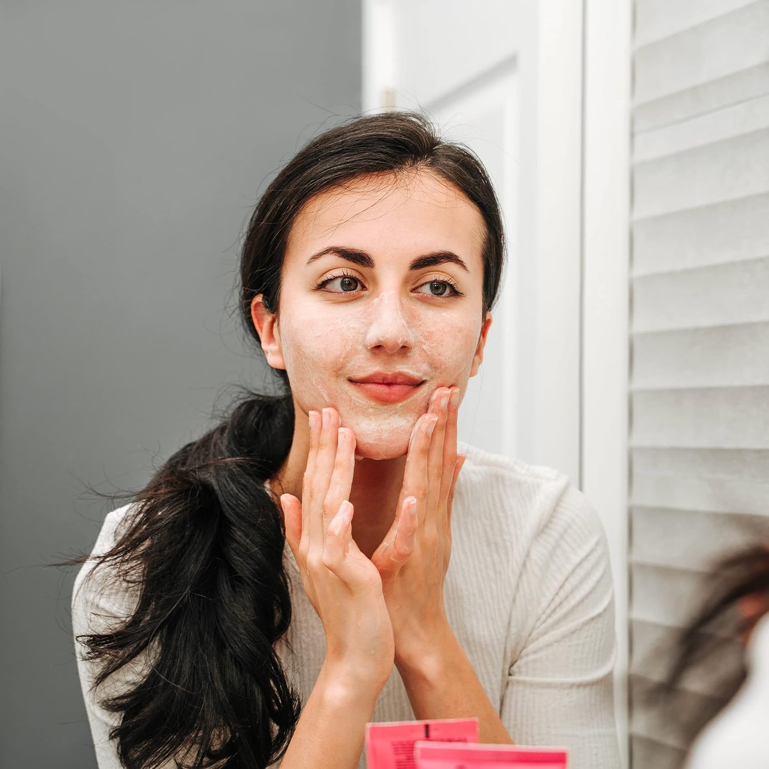Frau massiert AHA/PHA Gesichtsreiniger ins Gesicht vor Spiegel. Helle Geltextur auf Haut sichtbar. Produktverpackung unten im Bild.