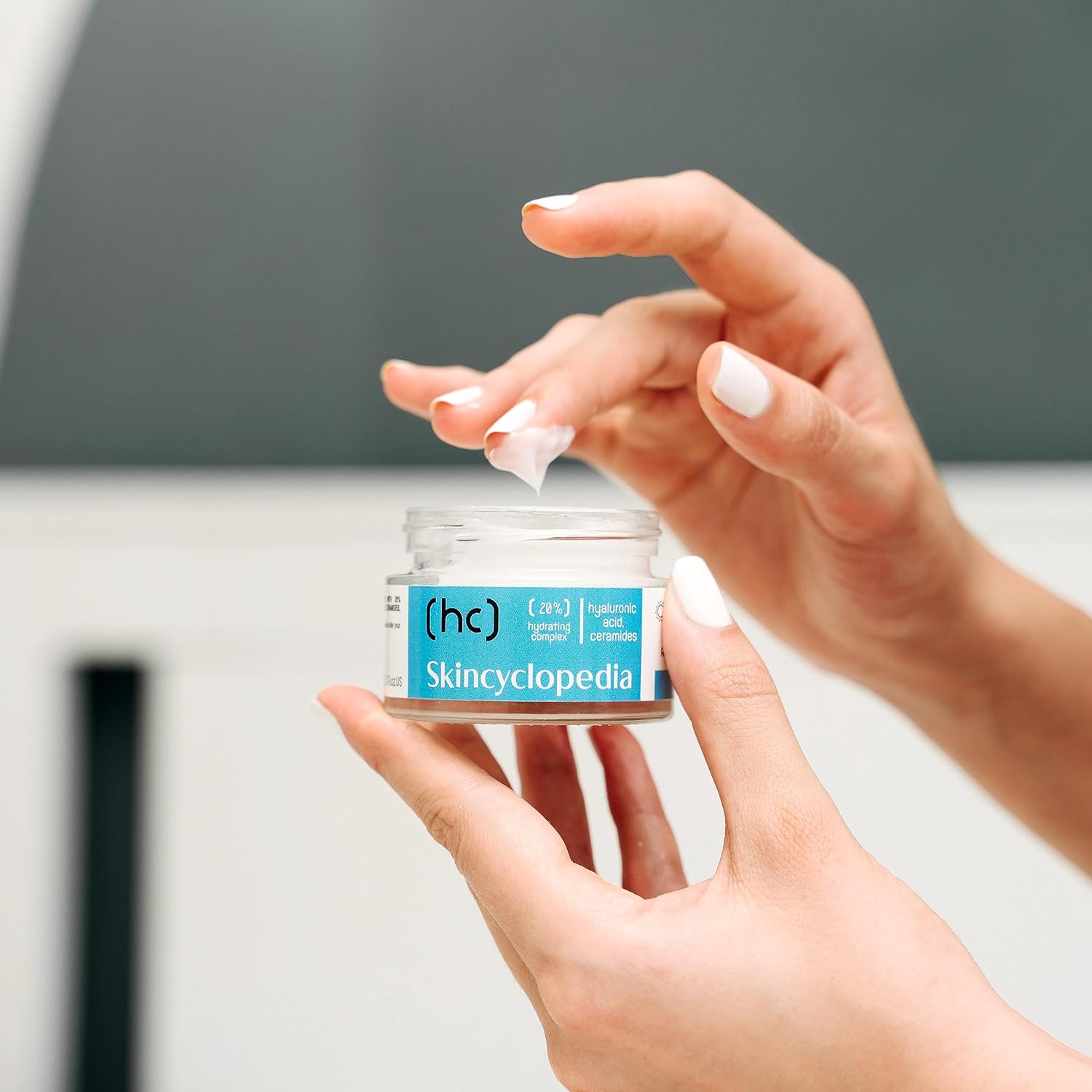 Hände halten eine SKINCYCLOPEDIA Feuchtigkeitscreme. Eine Fingerspitze berührt die Creme. Fokus auf Produktlabel.