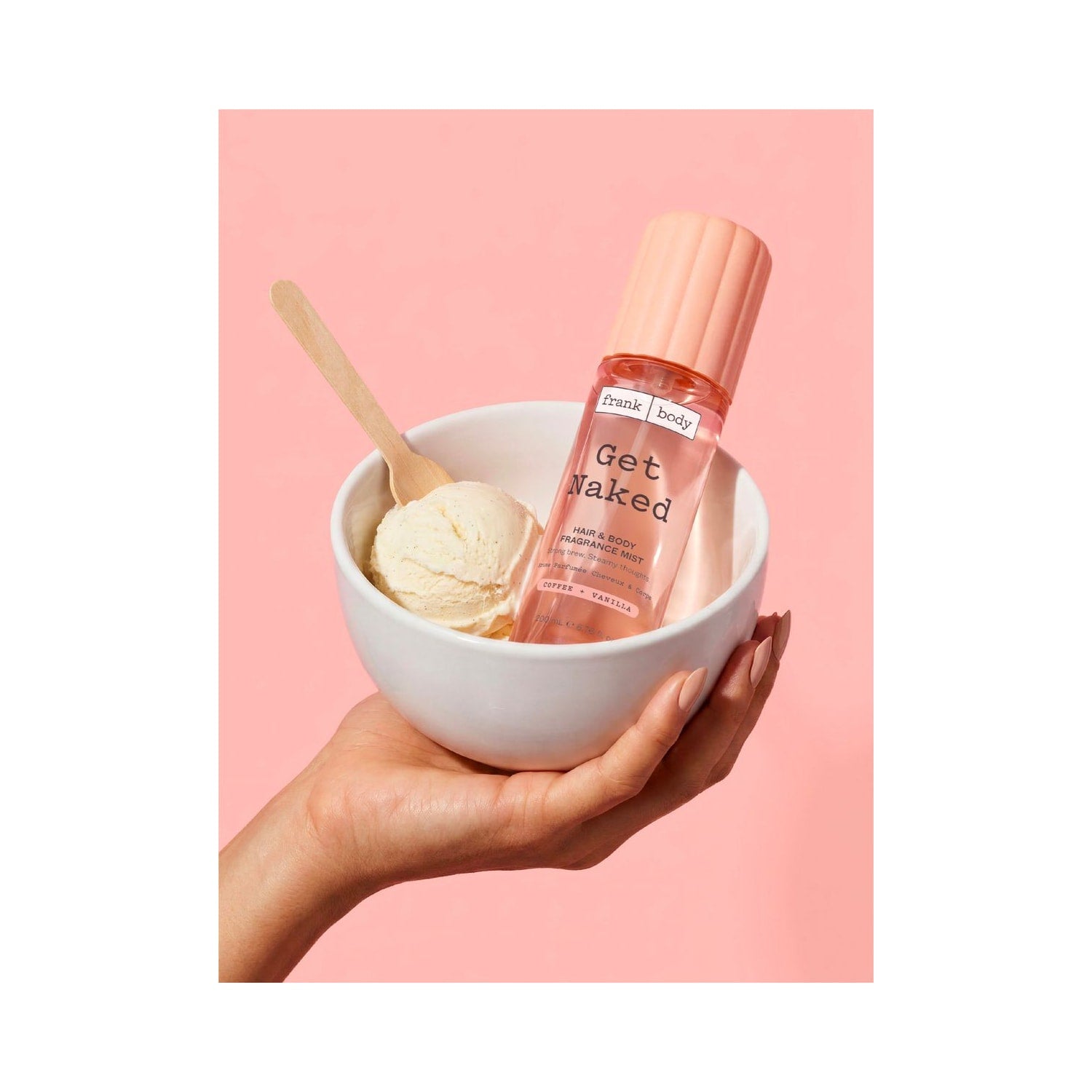 Una mano regge una coppetta di gelato alla vaniglia e il FRANK BODY Get Naked Hair & Body Fragrance Mist su uno sfondo rosa