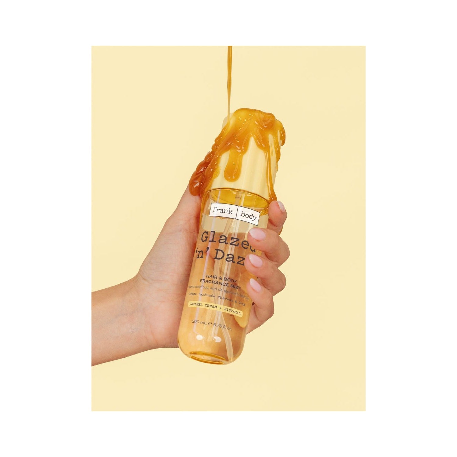 Una mano regge il profumo spray per capelli e corpo FRANK BODY Glazed 'n' Dazed, mentre del caramello liquido vi scorre sopra
