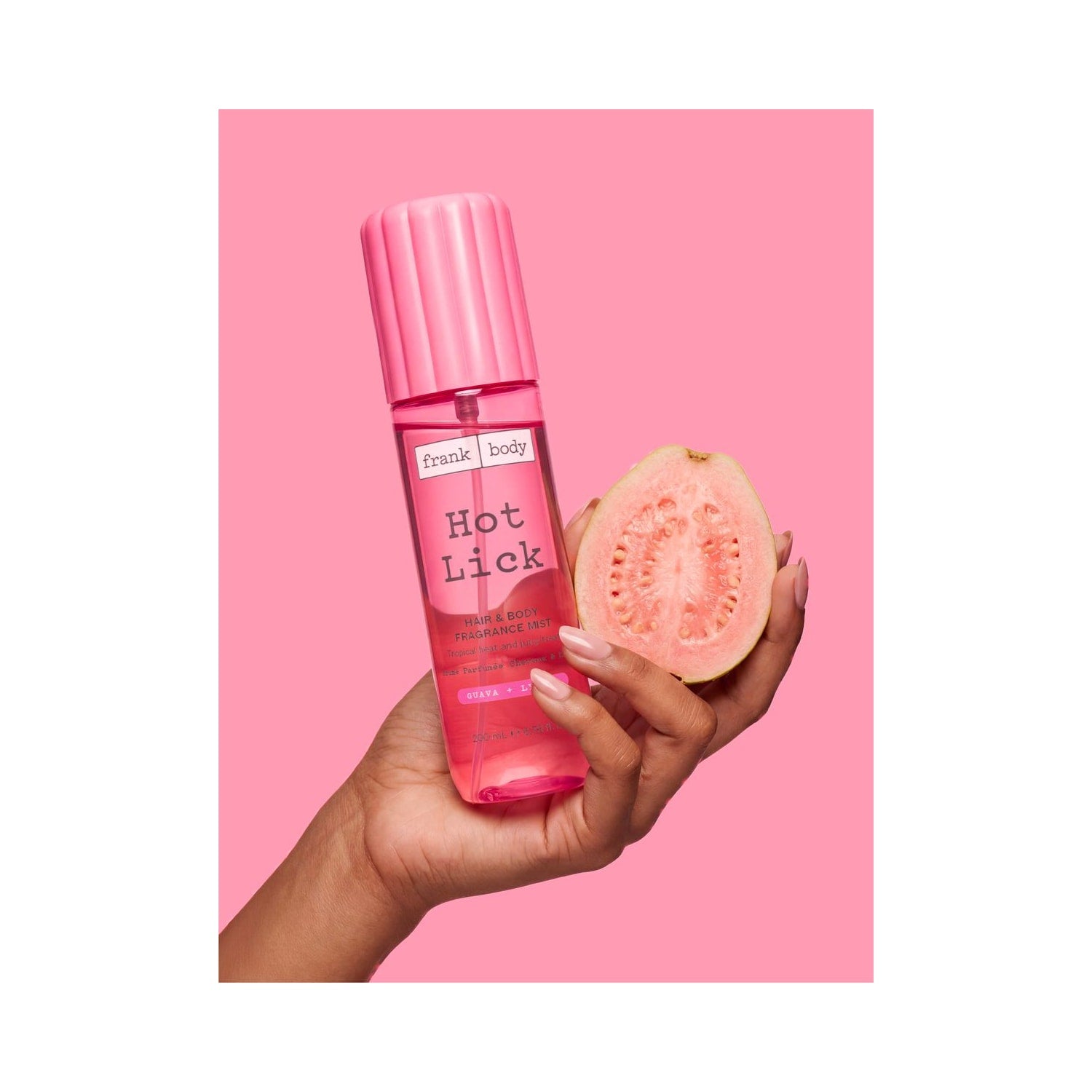Una mano regge il profumo per capelli e corpo FRANK BODY Hot Lick Hair & Body Fragrance Mist, di colore rosa, e mezza guava su uno sfondo rosa