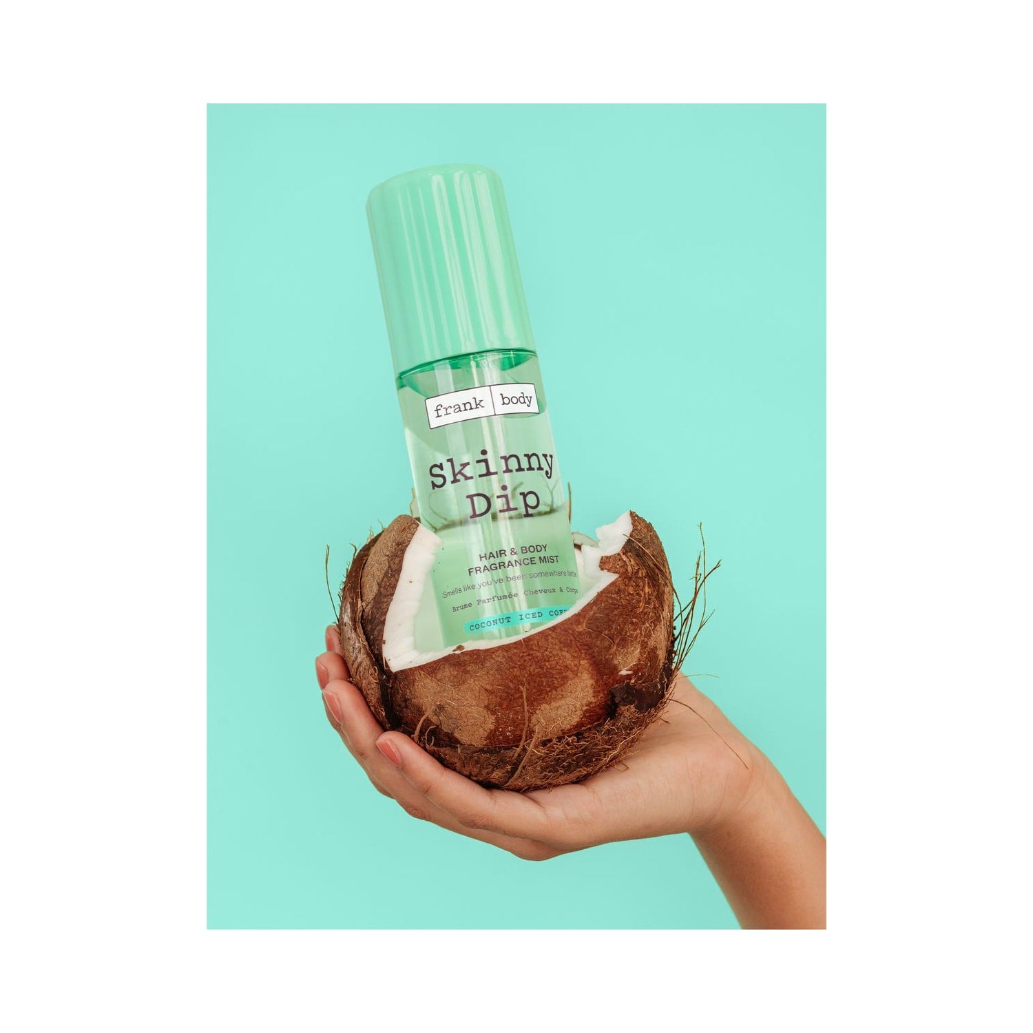 Una mano regge il FRANK BODY Skinny Dip Hair & Body Fragrance Mist all'interno di una noce di cocco su uno sfondo turchese