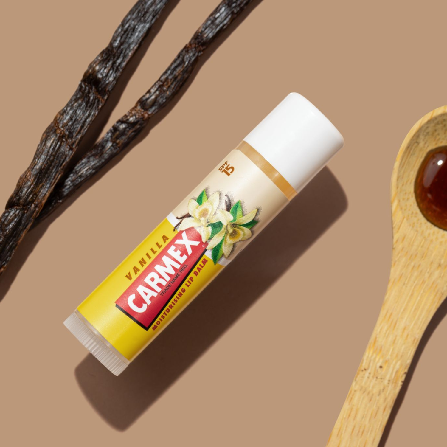 Ein Carmex Vanilla Lippenbalsam-Stick liegt mit Vanilleschoten und einem Löffel mit Flüssigkeit auf braunem Grund.