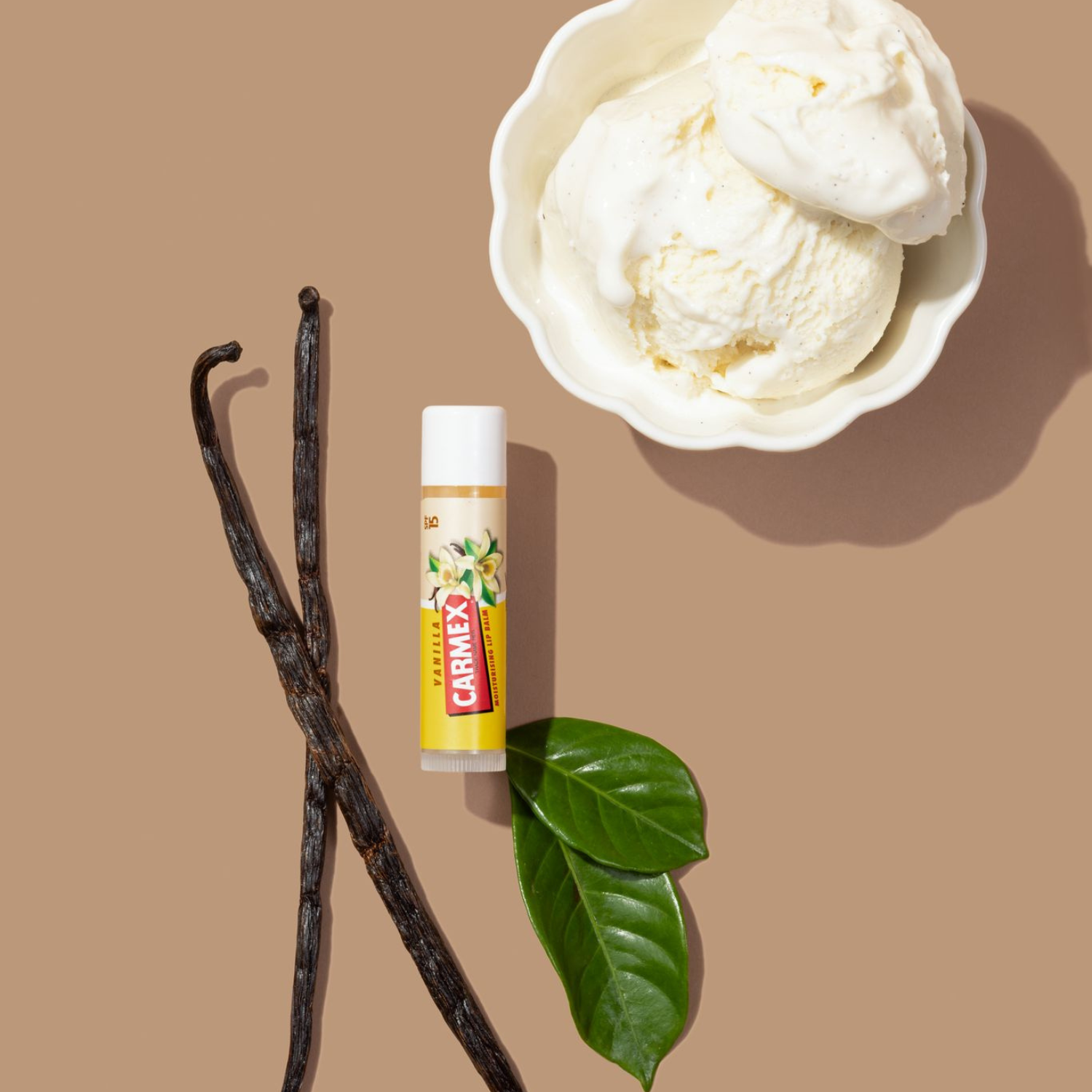 Carmex Vanilla Lippenbalsam Stick neben Vanilleeis, Vanilleschoten und grünen Blättern auf beigem Hintergrund.