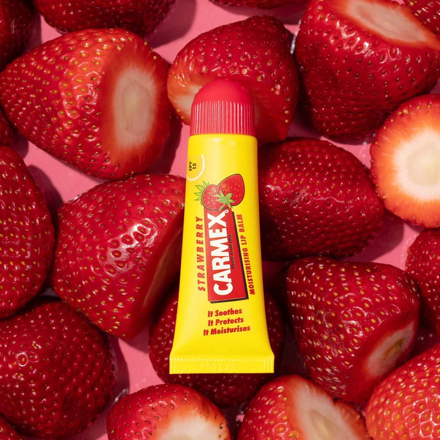 Gelbe Tube Carmex Erdbeer Lippenbalsam, umgeben von frischen Erdbeeren auf rosa Untergrund.