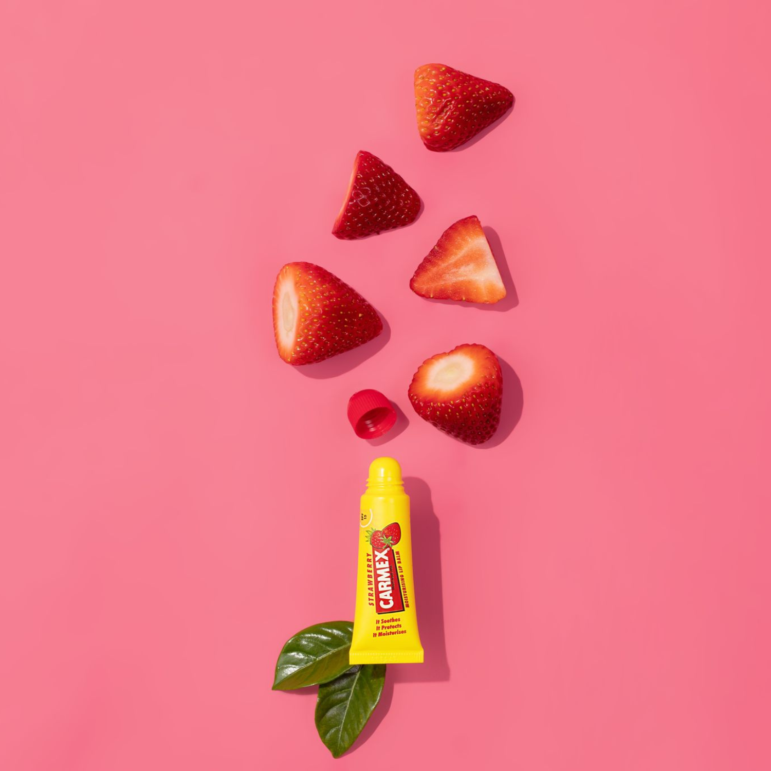 Gelbe Carmex Erdbeer Lippenbalsam Tube mit Erdbeerstücken und grünen Blättern auf pinkem Hintergrund.