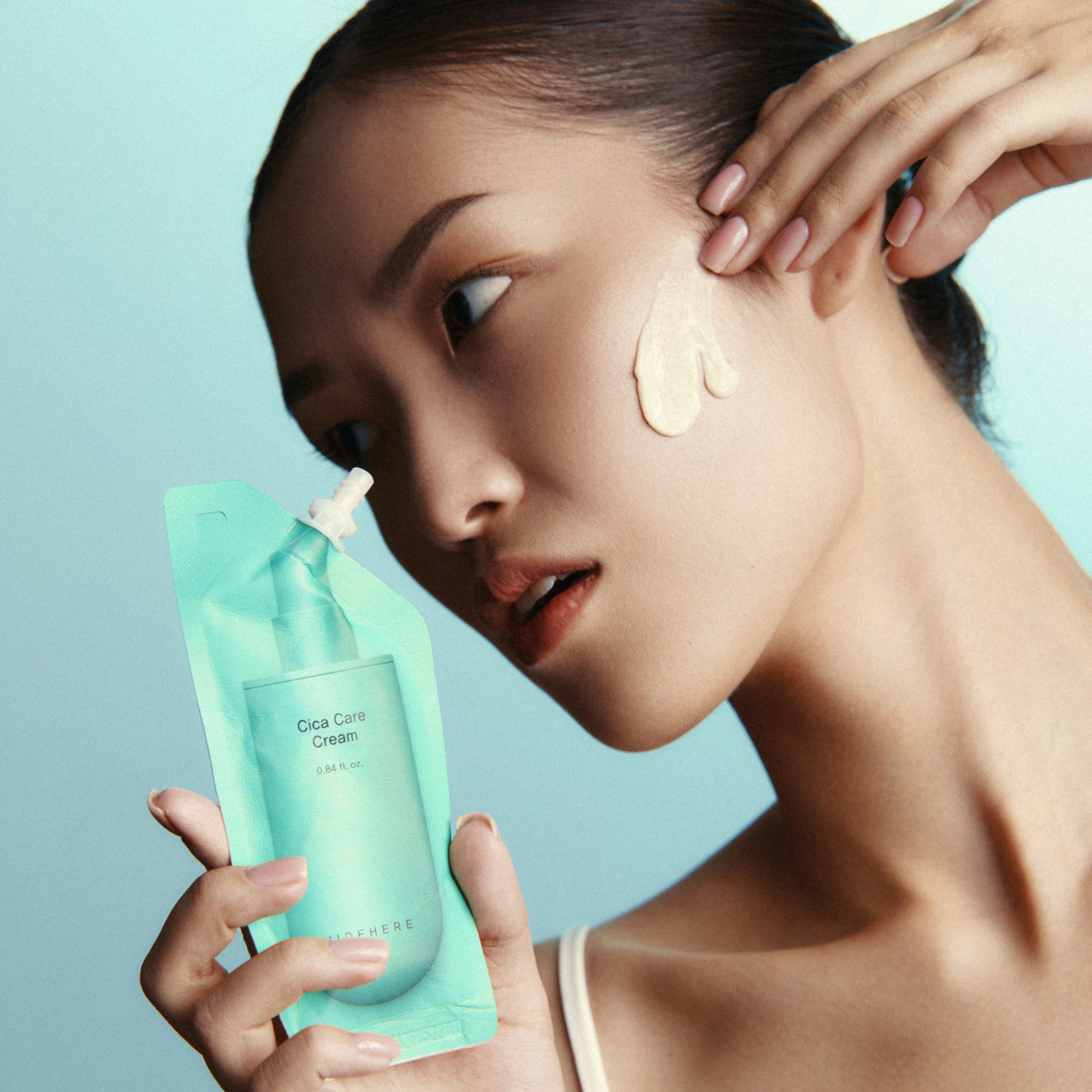Eine Person trägt Cica Care Cream auf ihr Gesicht auf. Sie hält den Produktbehälter in der einen Hand und tupft mit der anderen Hand die Creme auf ihre Wange, vor einem hellblauen Hintergrund.
