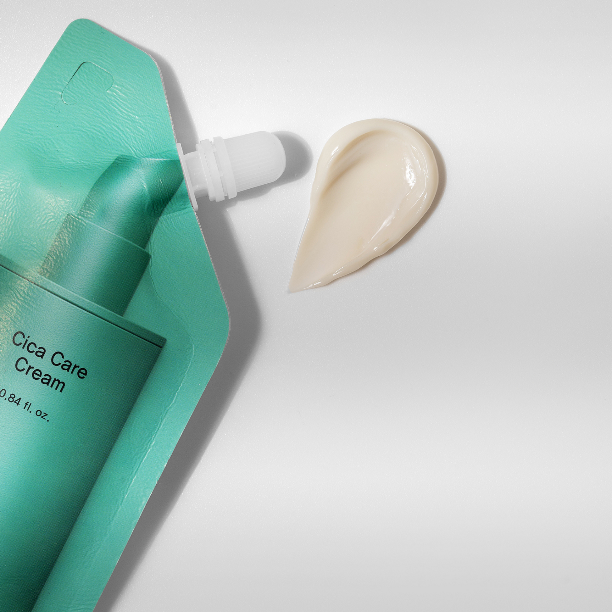 Eine tealfarbene Cica-Pflegecreme-Packung mit weißer Tülle liegt neben einem tropfenförmigen Cremestrich auf einer weißen Fläche.