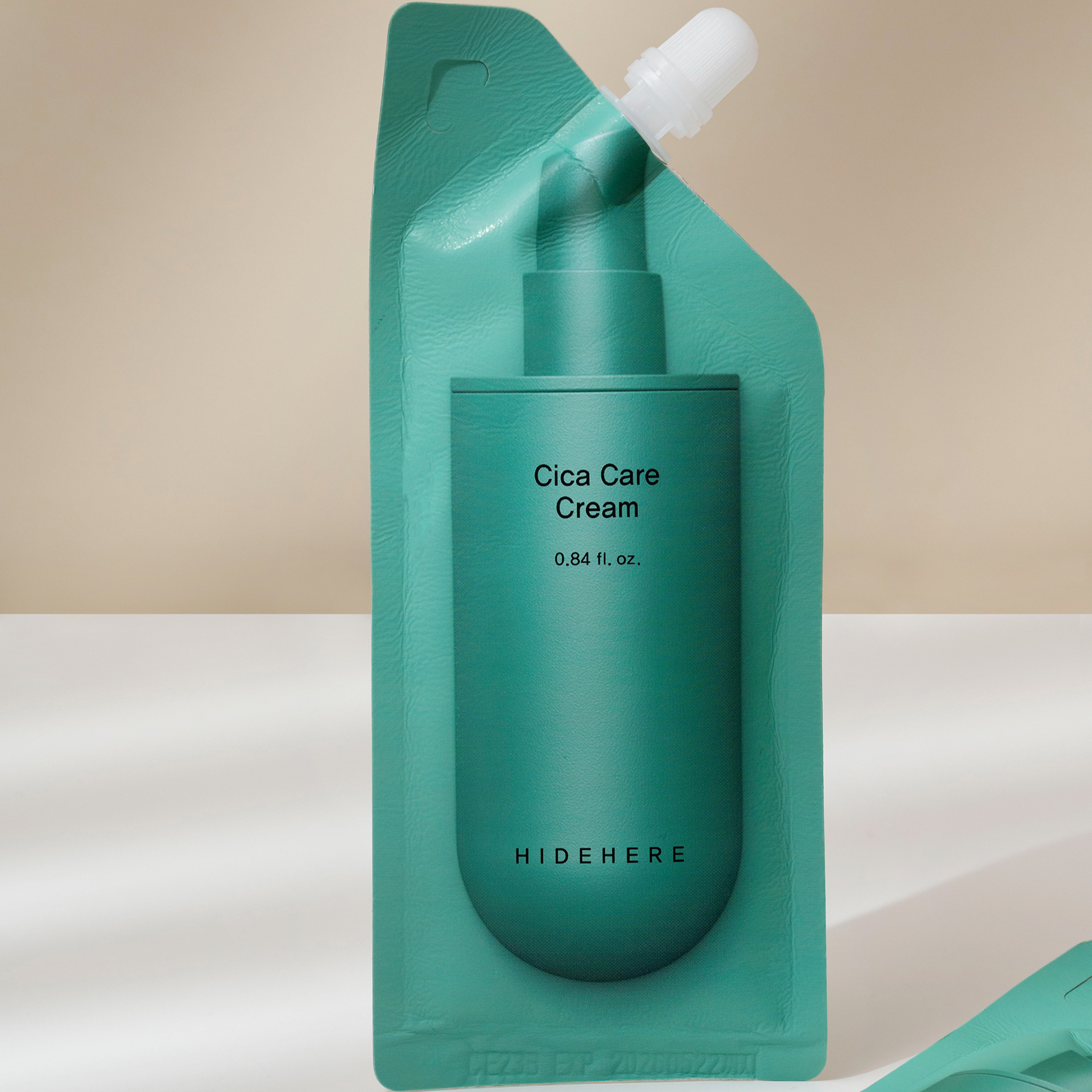 Ein blaugrüner Beutel mit Cica-Pflegecreme und weißer Kappe steht mittig auf einer weißen Fläche. Darauf steht "0.84 fl. oz.". Der Markenname "HIDEHERE" ist unten zu sehen. Die Creme ist vor einem hellbeigen Hintergrund abgebildet.