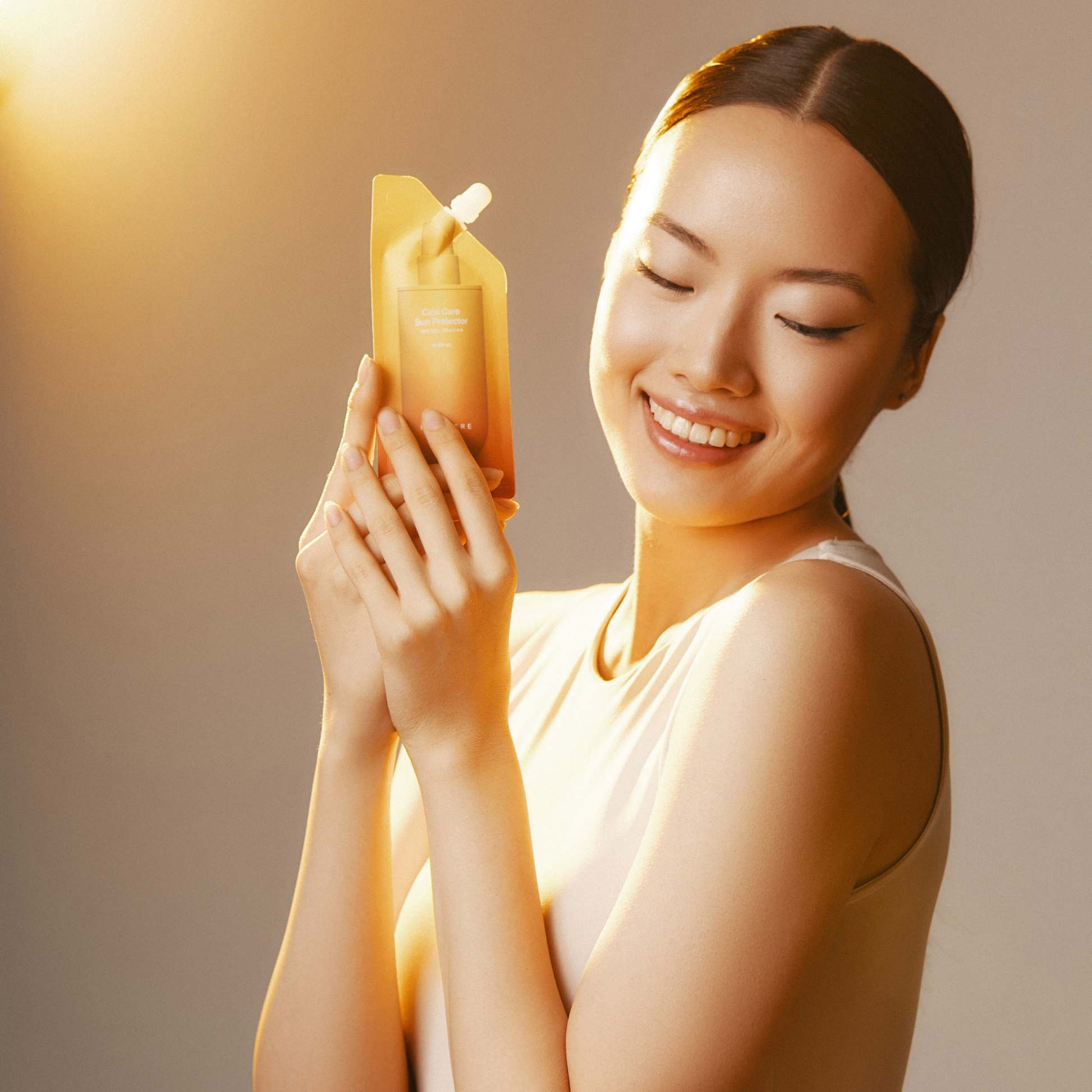Eine lächelnde Asiatin hält ein gelbes Sonnenschutzpaket in der Hand. Sie hat helle Haut, dunkles Haar, das nach hinten gezogen ist, und trägt ein weißes Oberteil. Von links scheint ein Licht, das einen warmen Schein wirft.