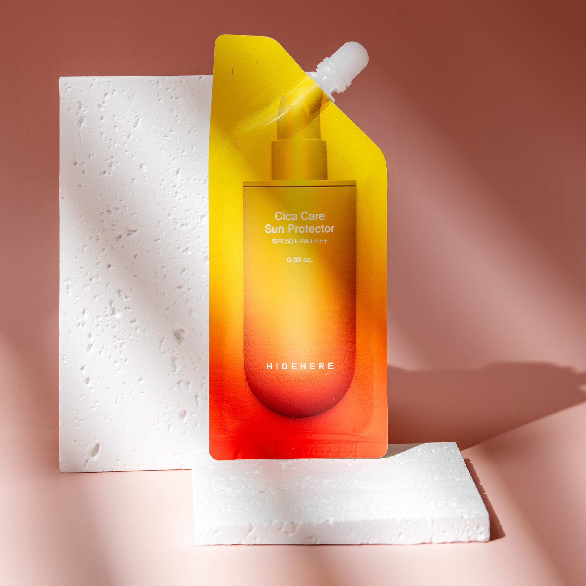 Produktfoto von Cica Care Sun Protector in einem farbenfrohen gelb-roten Beutel mit weißem Spenderaufsatz, präsentiert auf strukturierten weißen Blöcken vor einem zartrosa Hintergrund.