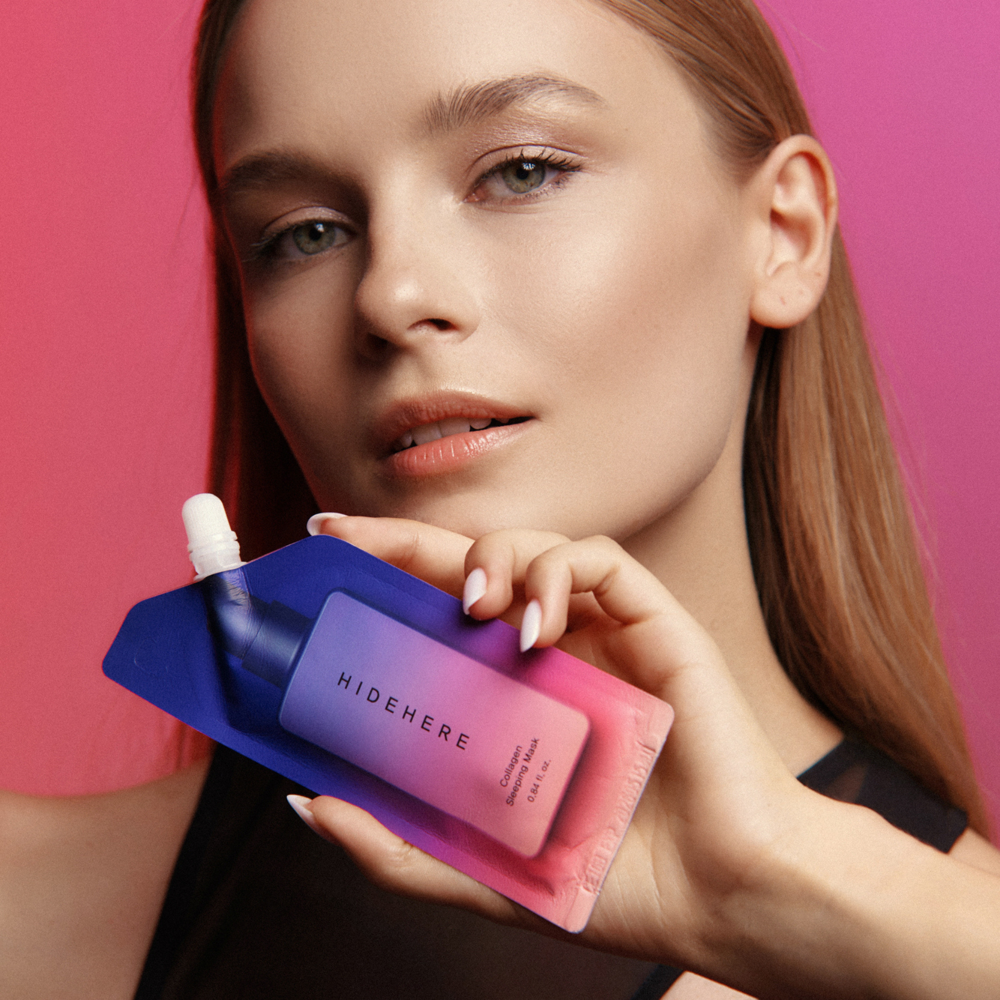 Eine Frau hält eine "HIDEHERE"-Kollagen-Schlafmaske vor einem Hintergrund mit rosa und magentafarbenem Farbverlauf. Die Produktverpackung ist lila, rosa und blau. Ihr Haar ist blond und glatt.
