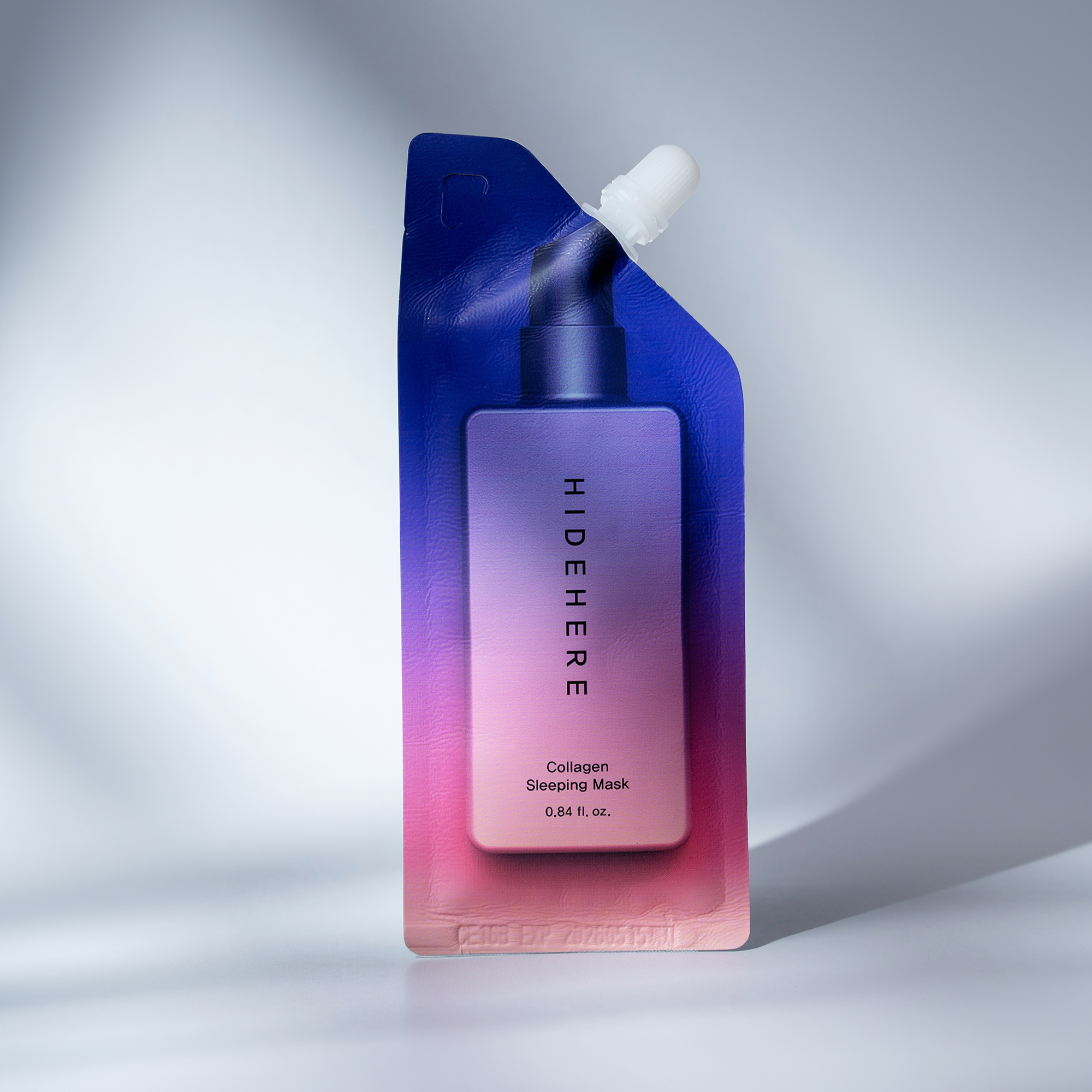 Ein Beutel mit der Kollagen-Schlafmaske "Hide Here", deren Farbe von Rosa zu Blau übergeht, steht vor einem weißen Hintergrund mit Schatten. Eine weiße Kappe befindet sich auf der Oberseite.