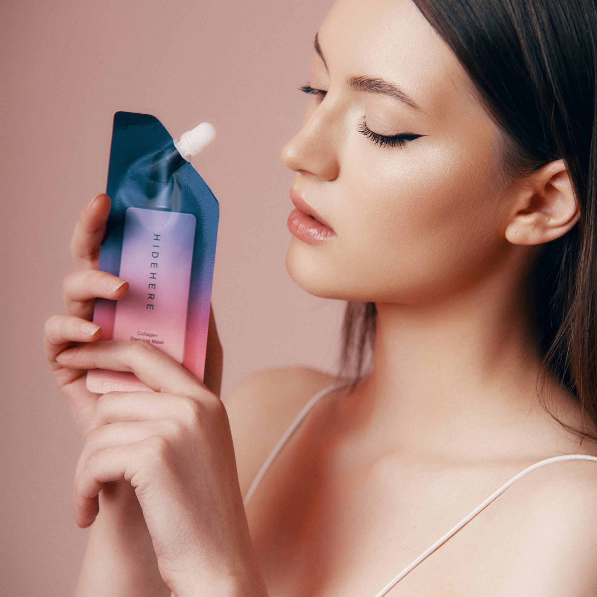 Eine Frau in einem weißen Oberteil hält einen Beutel mit der Collagen-Schlafmaske "HideHere" in der Hand. Sie hat dunkles Haar, helle Haut und schaut mit geschlossenen Augen auf den Beutel. Der Hintergrund ist ein zartes Rosa.
