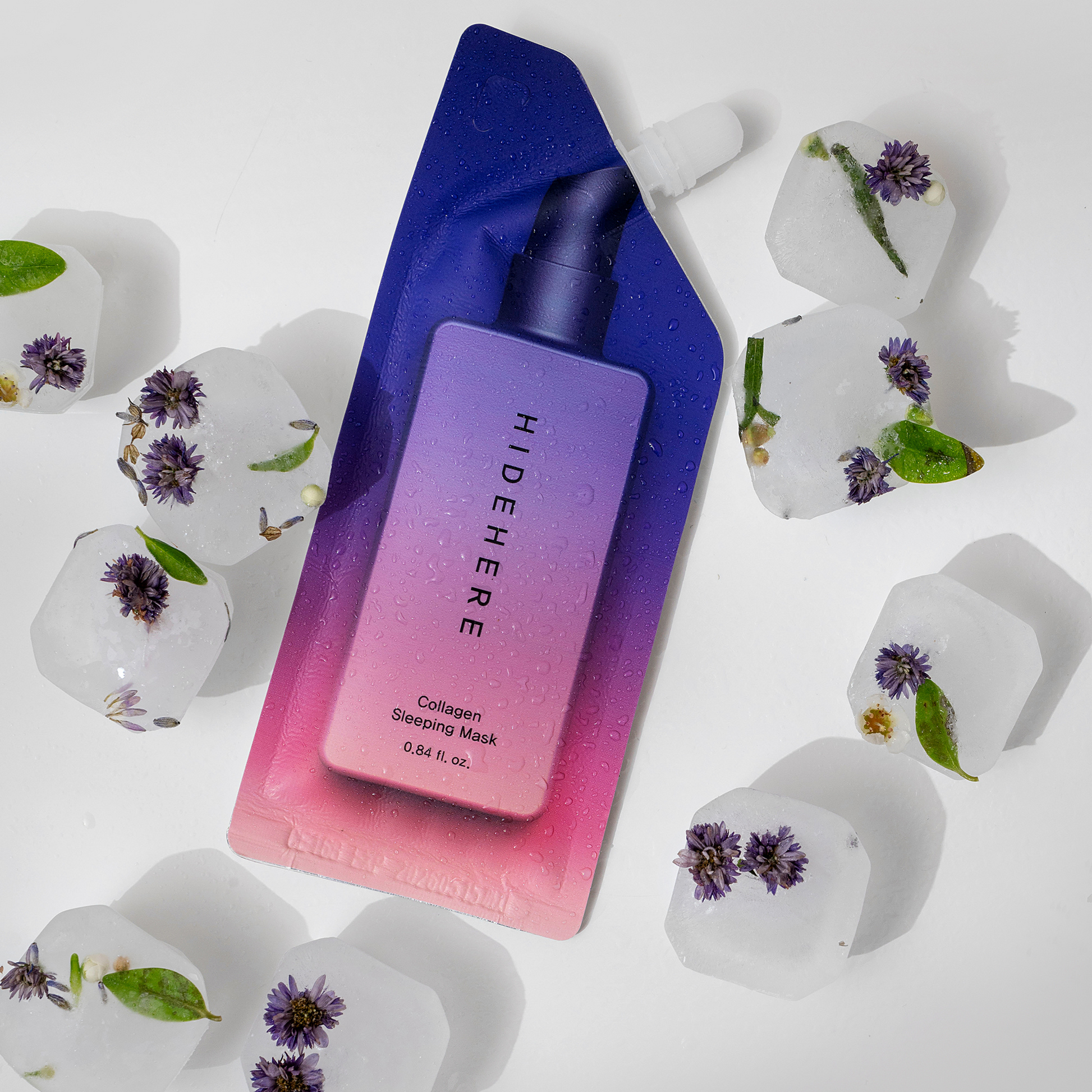 Eine Packung der HideHere Collagen Sleeping Mask auf weißem Hintergrund, umgeben von Eiswürfeln mit eingefrorenen Blumen darin. Die Verpackung hat einen blau-lila-rosa Farbverlauf.