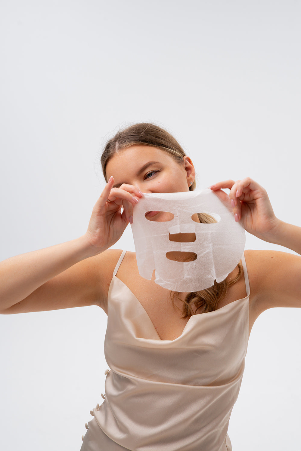 Frau hält Polyglutaminsäure Sheet Maske vor dem Gesicht. Die Tuchmaske hat Aussparungen für Augen, Nase und Mund. Sie trägt ein helles Top.