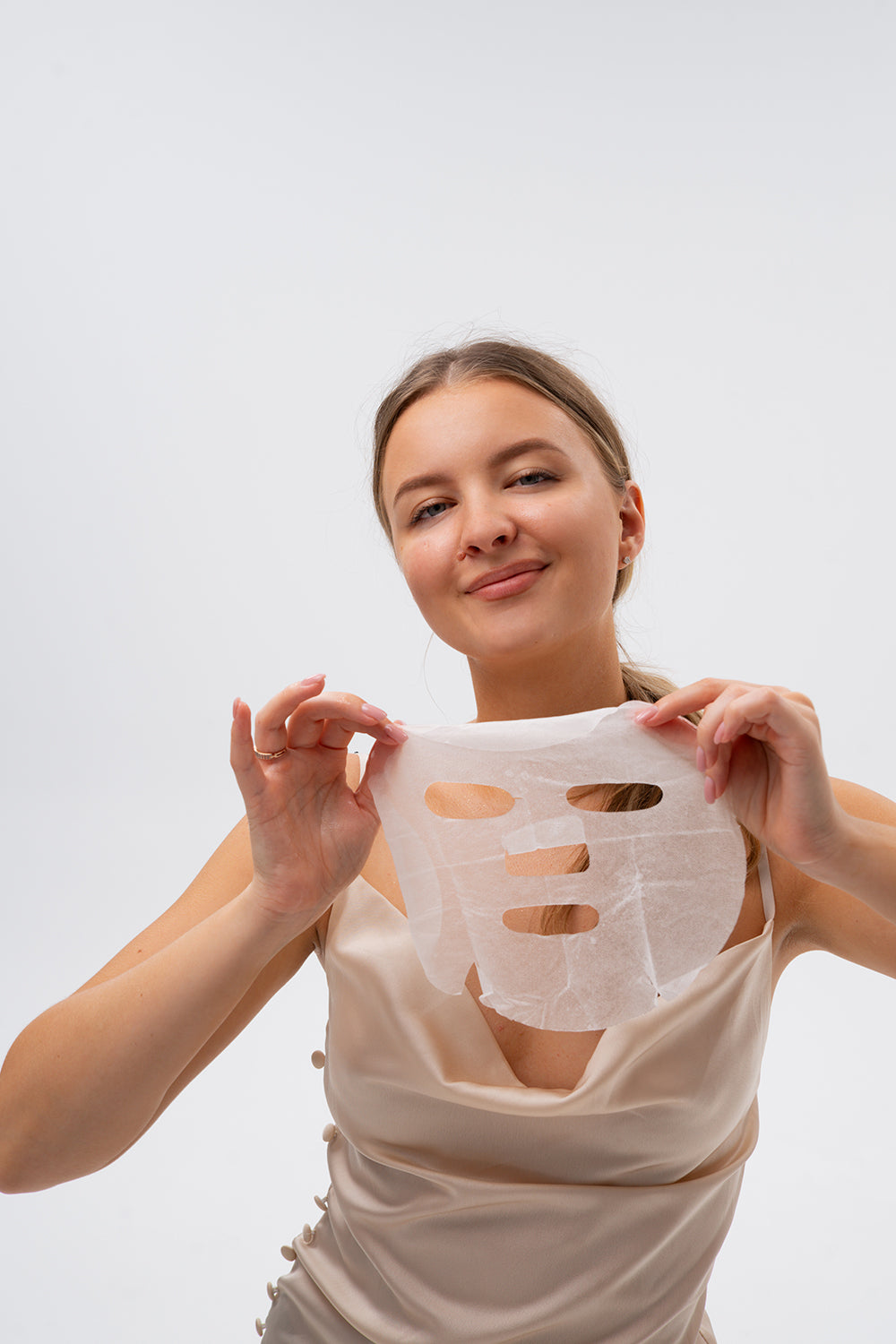 Eine Frau hält eine weiße Tuchmaske mit Aussparungen für Augen und Mund vor ihr Gesicht. Sie hat ein helles Top an und lächelt.
