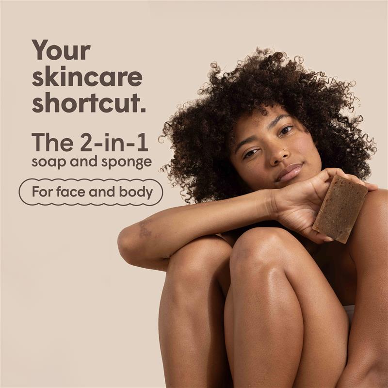 Eine Frau mit Afro-Haar hält eine rechteckige Seife in der Hand, während sie auf einem beigen Hintergrund sitzt. Text: "Your skincare shortcut. The 2-in-1 soap and sponge. For face and body".